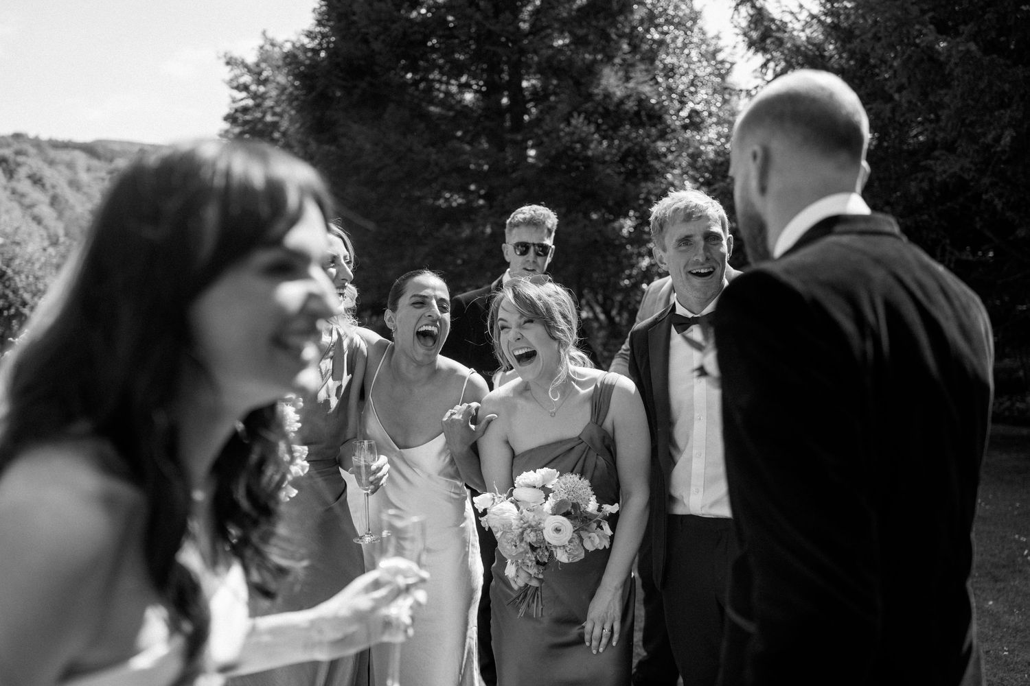 Black and white candid photo of wedding party laughing together during outdoor celebration.