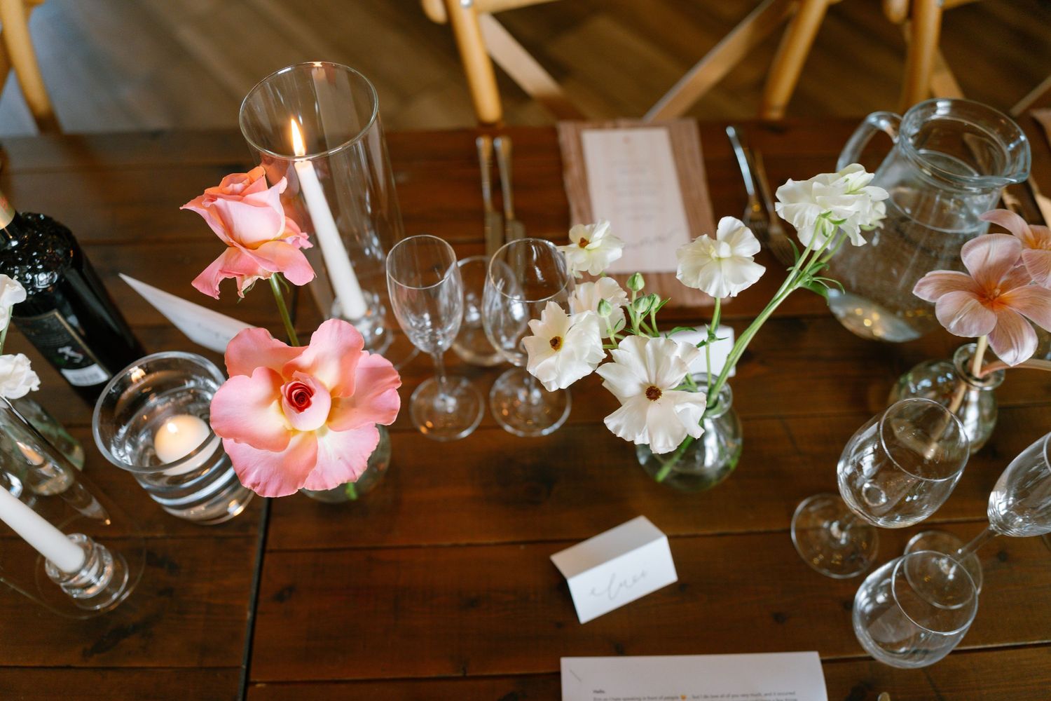 Wedding reception table decorated with pink flowers, glassware, and place cards on dark wood surface.