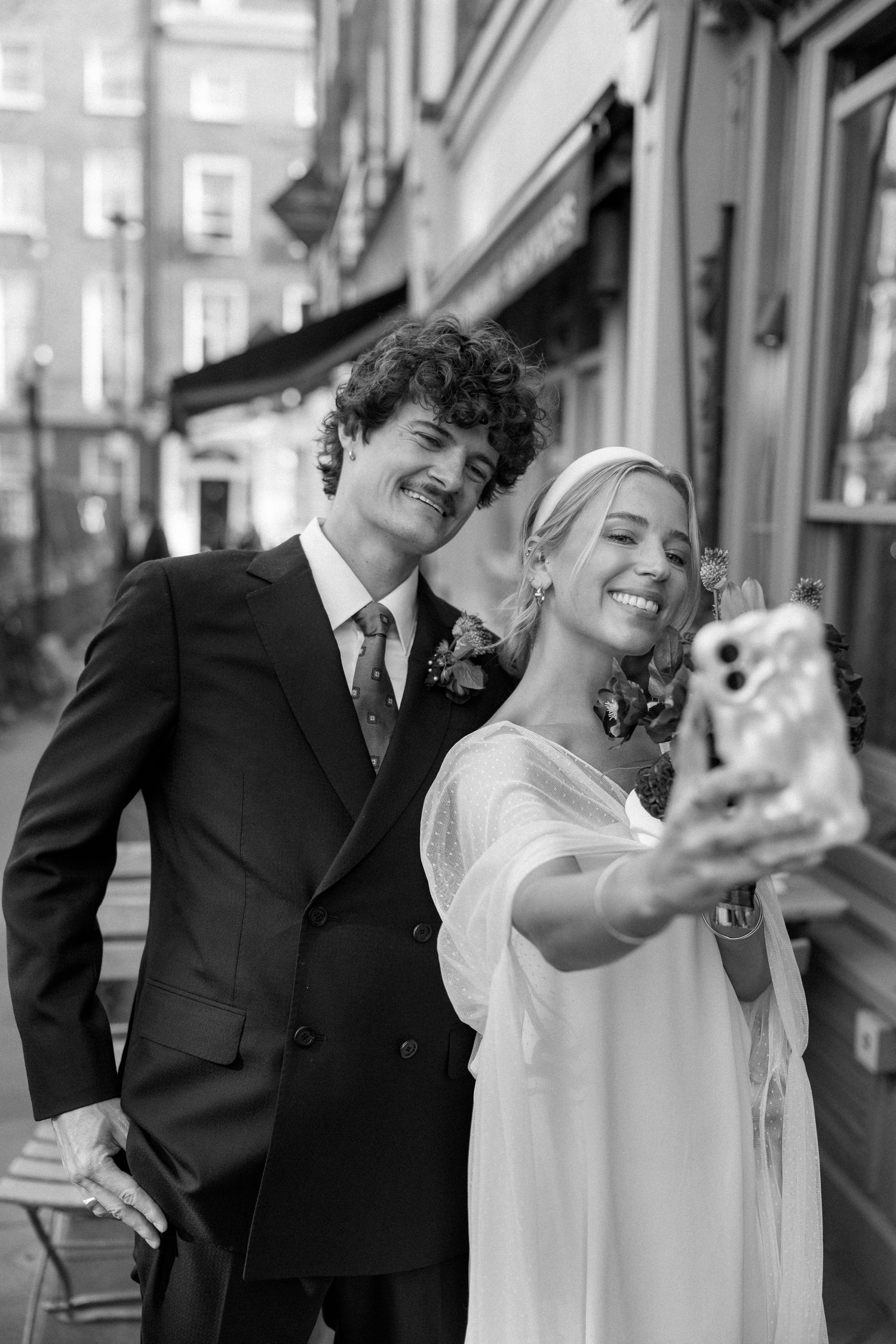 A wedding couple poses playfully for selfies outside a building on their special day.