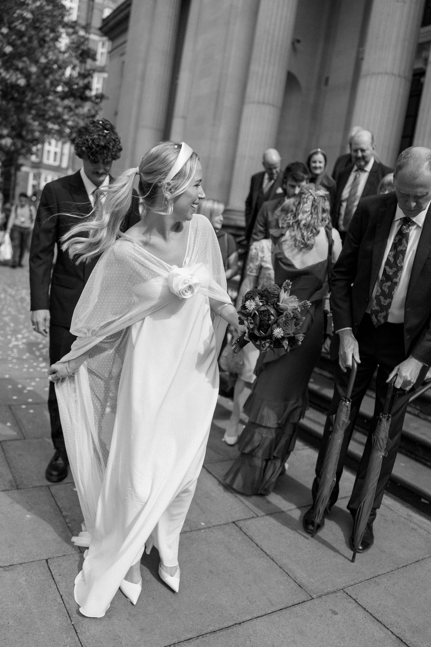 Bride in flowing white dress descends stone steps while guests look on.