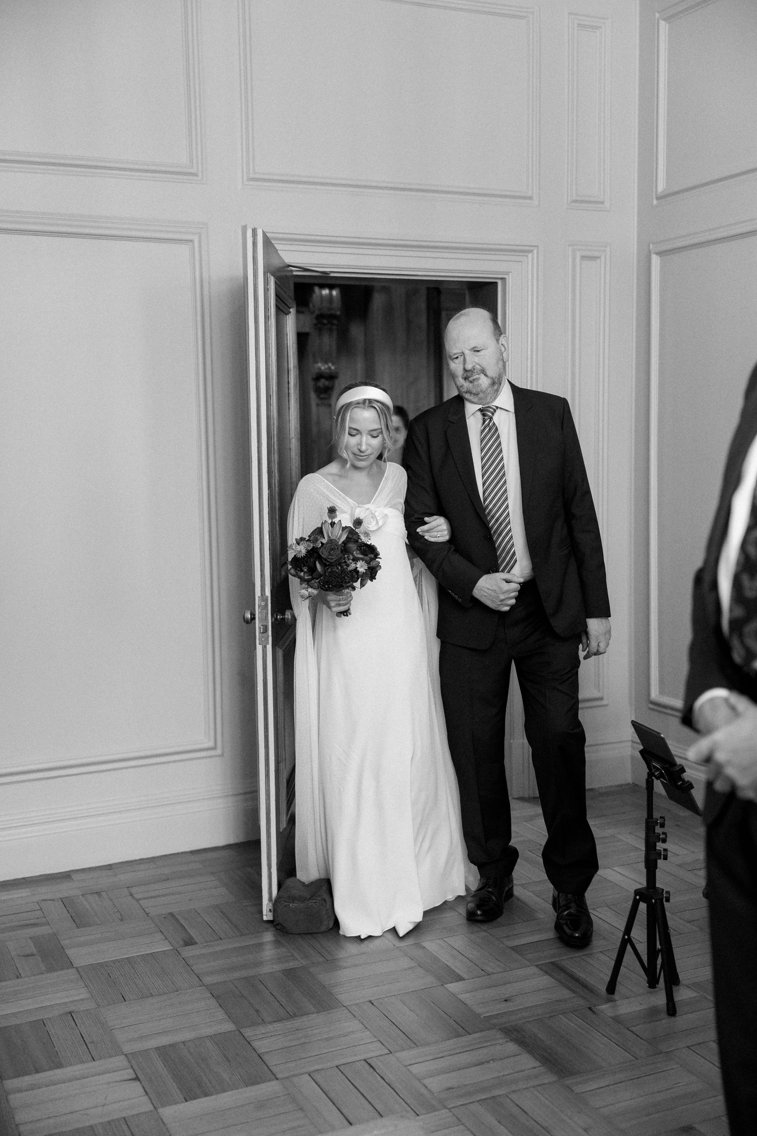 Wedding processional moment captured in black and white at elegant venue entrance.