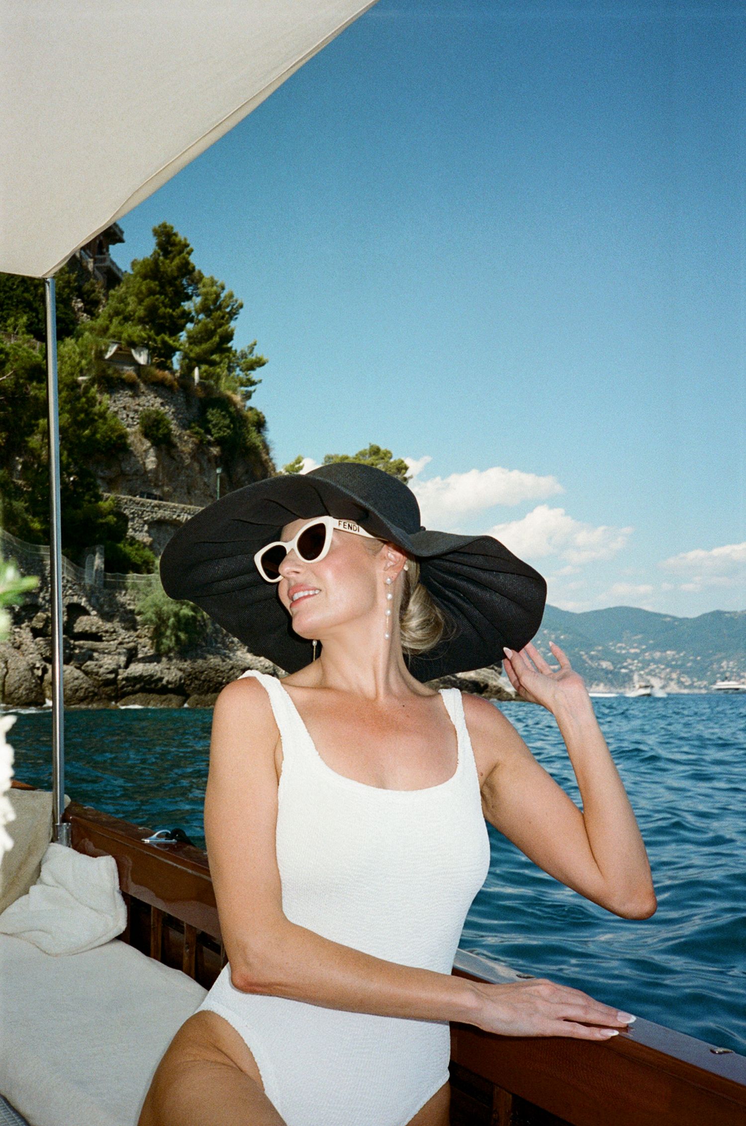 Woman in white swimsuit and black wide-brim hat on a luxury boat with blue Mediterranean sea backdrop.