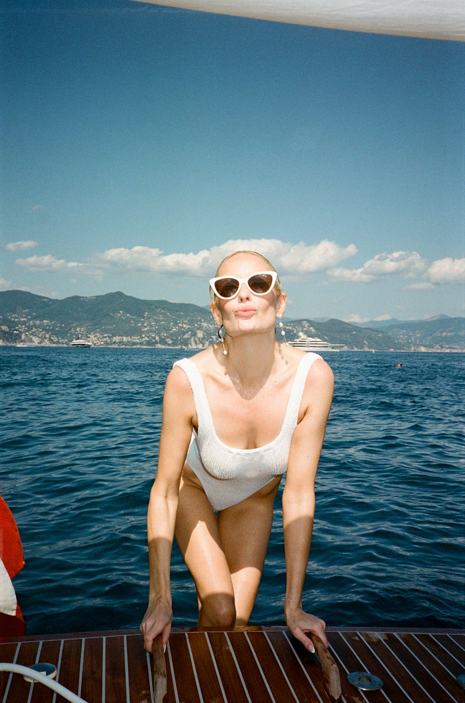 Woman in white swimsuit and cat-eye sunglasses posing on a yacht deck with Mediterranean sea and mountains behind.