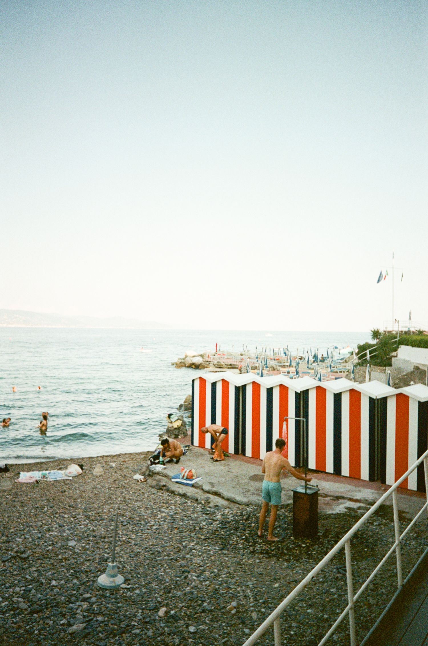 Striped red and white beach cabanas on a rocky Mediterranean shore with swimmers and a clear sky.