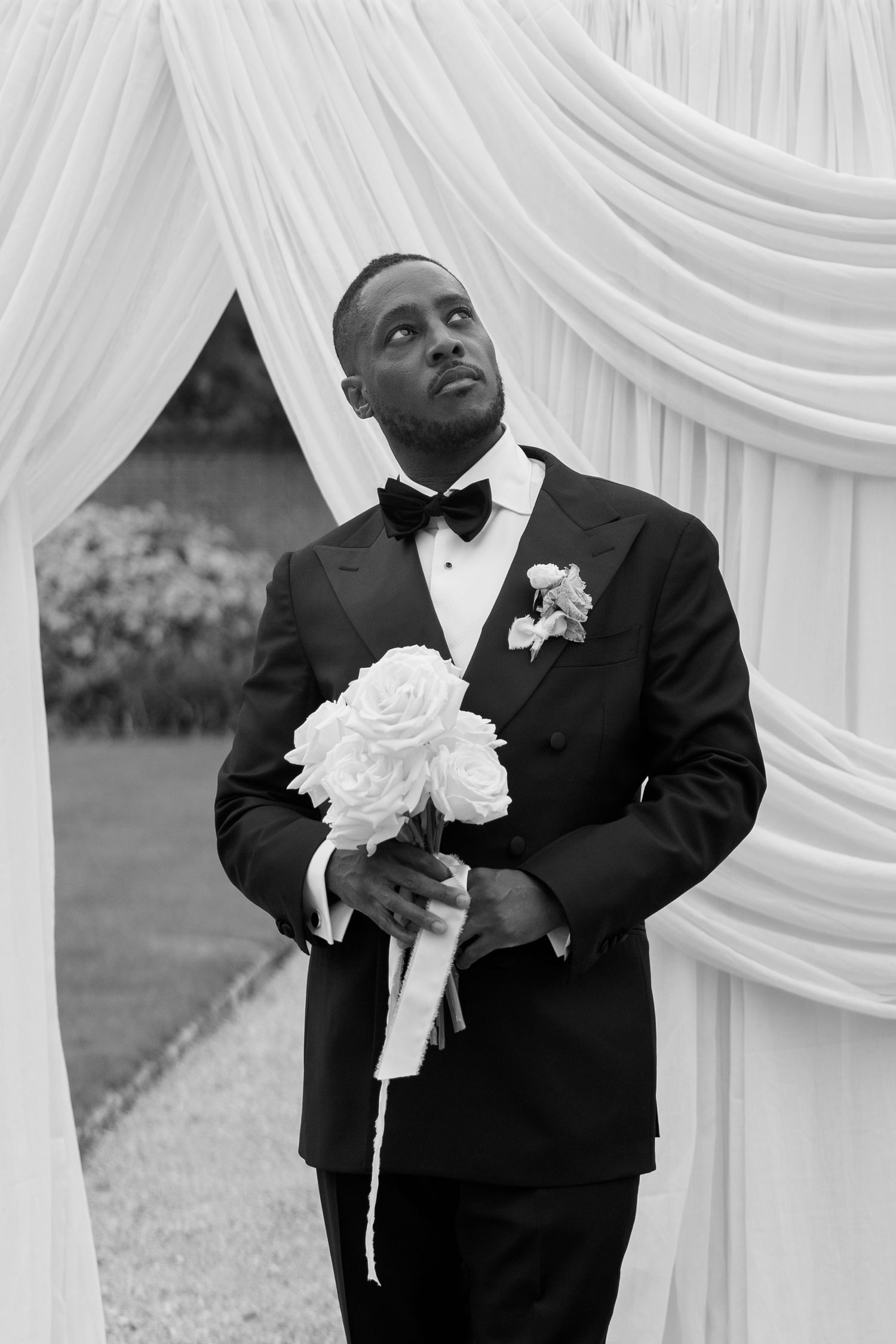 Black and white portrait of groom in tuxedo holding white rose bouquet under draped arch.