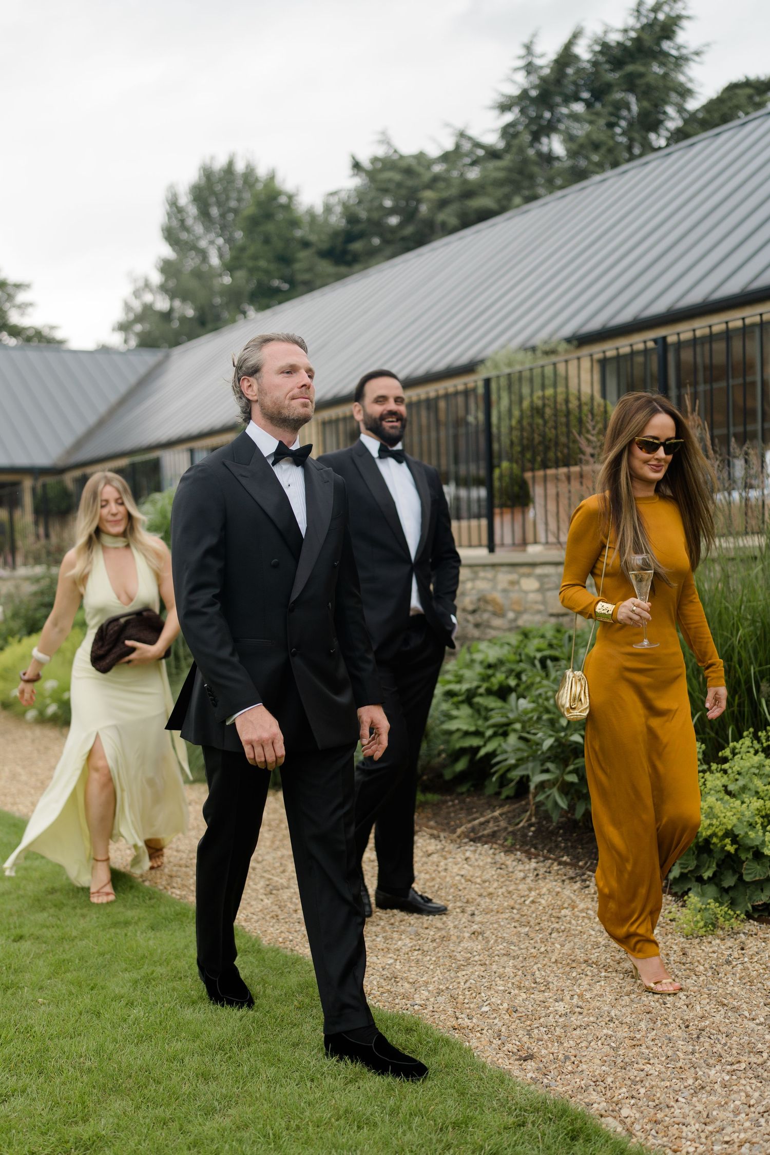 Wedding guests in formal attire walking along gravel path beside modern stone barn venue.