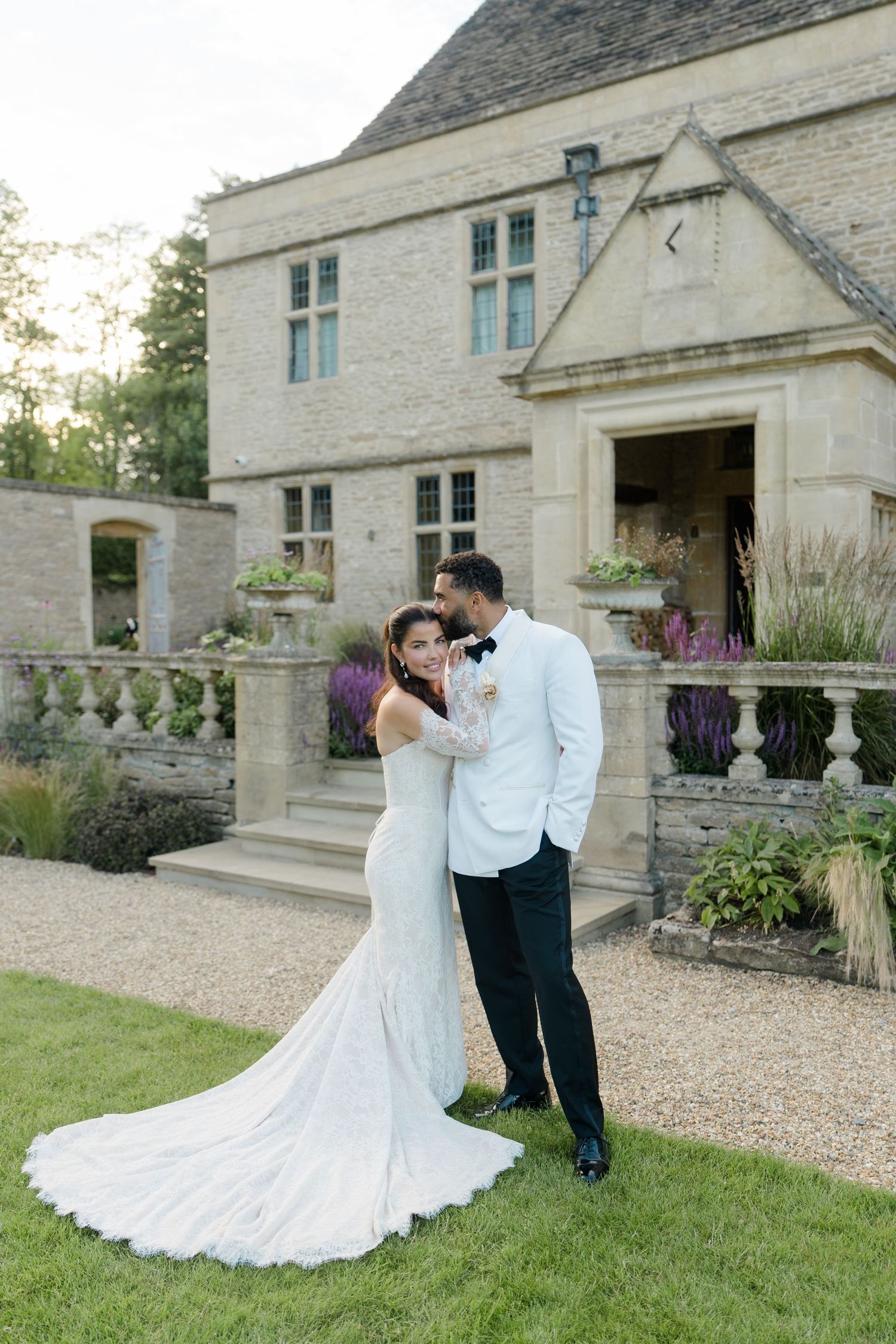 Couple embracing in wedding attire outside a grand stone English manor with lush garden landscaping.