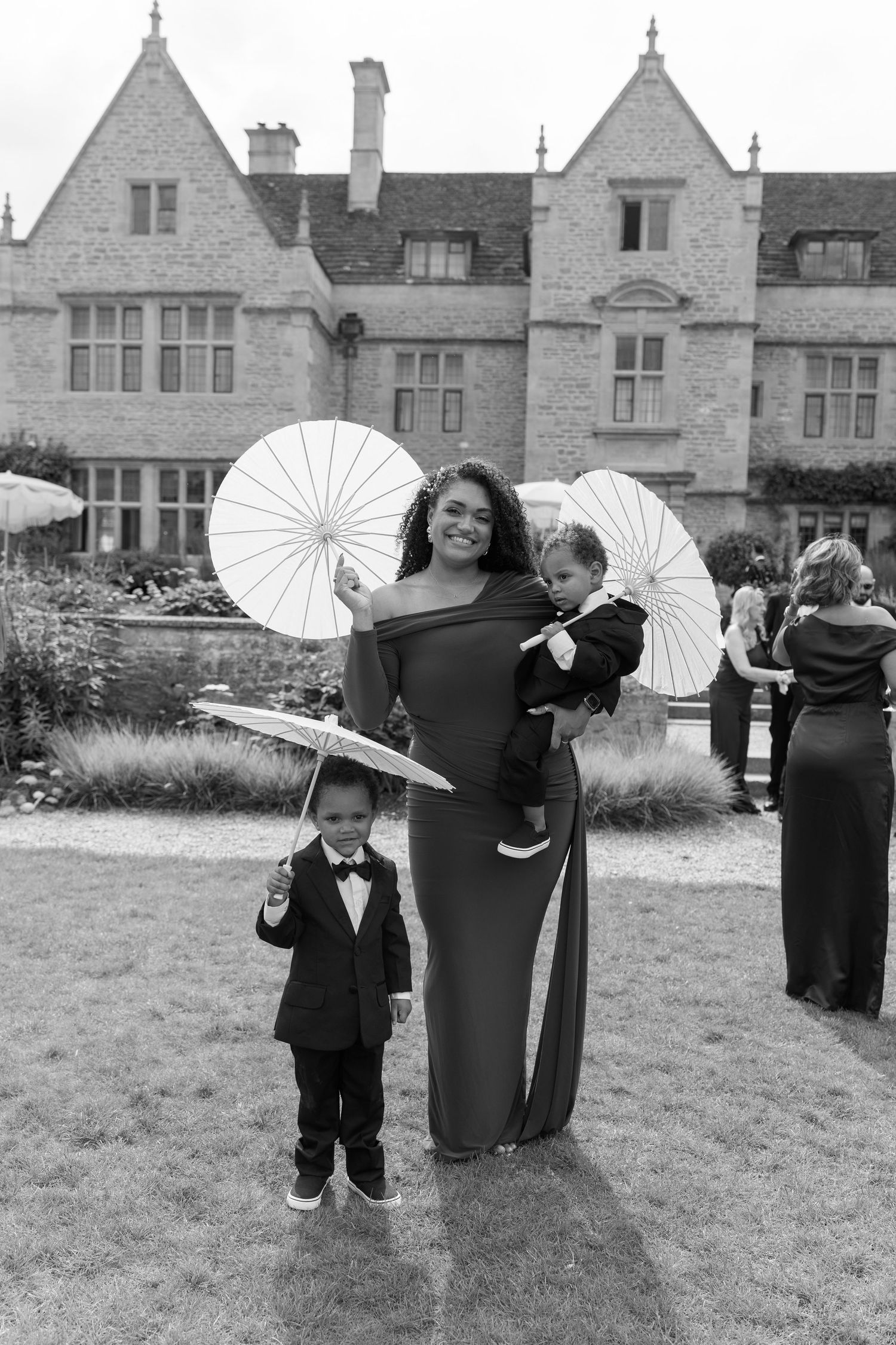 Woman in elegant gown holding baby and parasol beside young boy in tuxedo at wedding.