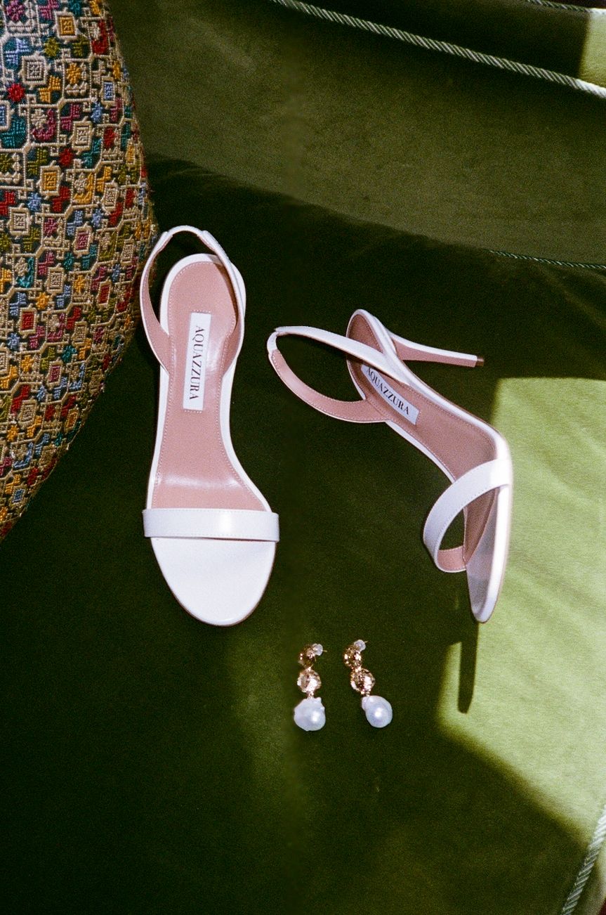 White strappy heeled sandals by Aquazzura displayed on green velvet beside delicate gold and pearl drop earrings.