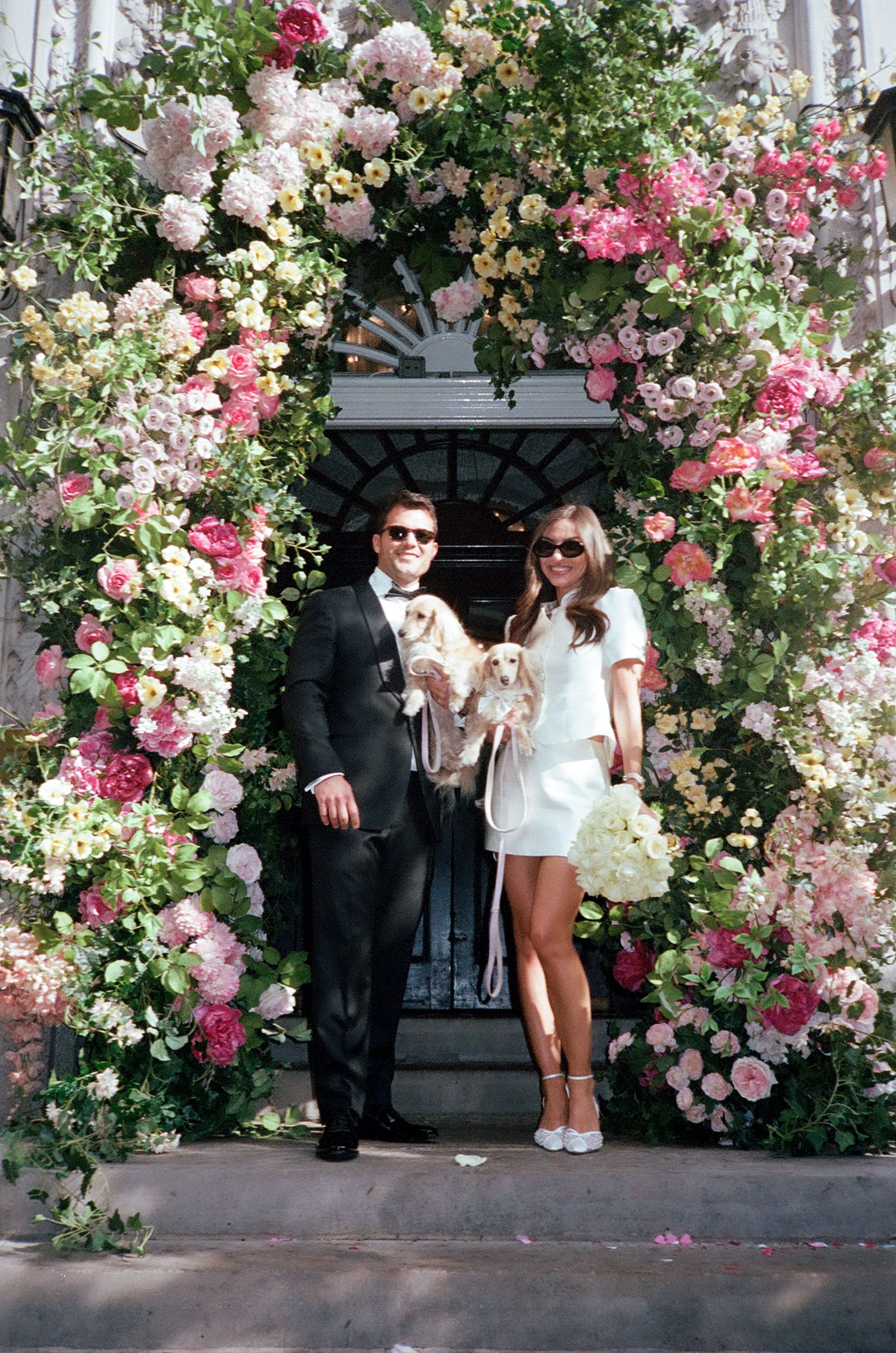 Couple in formal attire holding a dog under a lush floral arch with pink and white blooms at a wedding.
