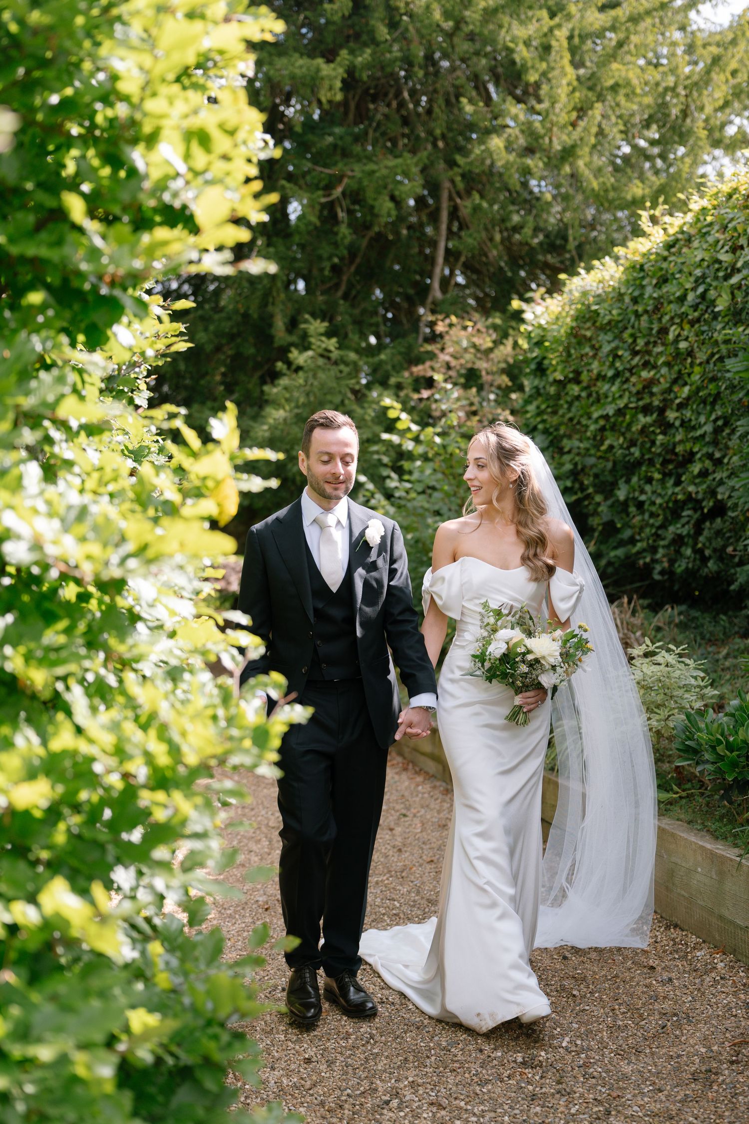 Couple walking along gravel garden path surrounded by green hedges on sunny day.