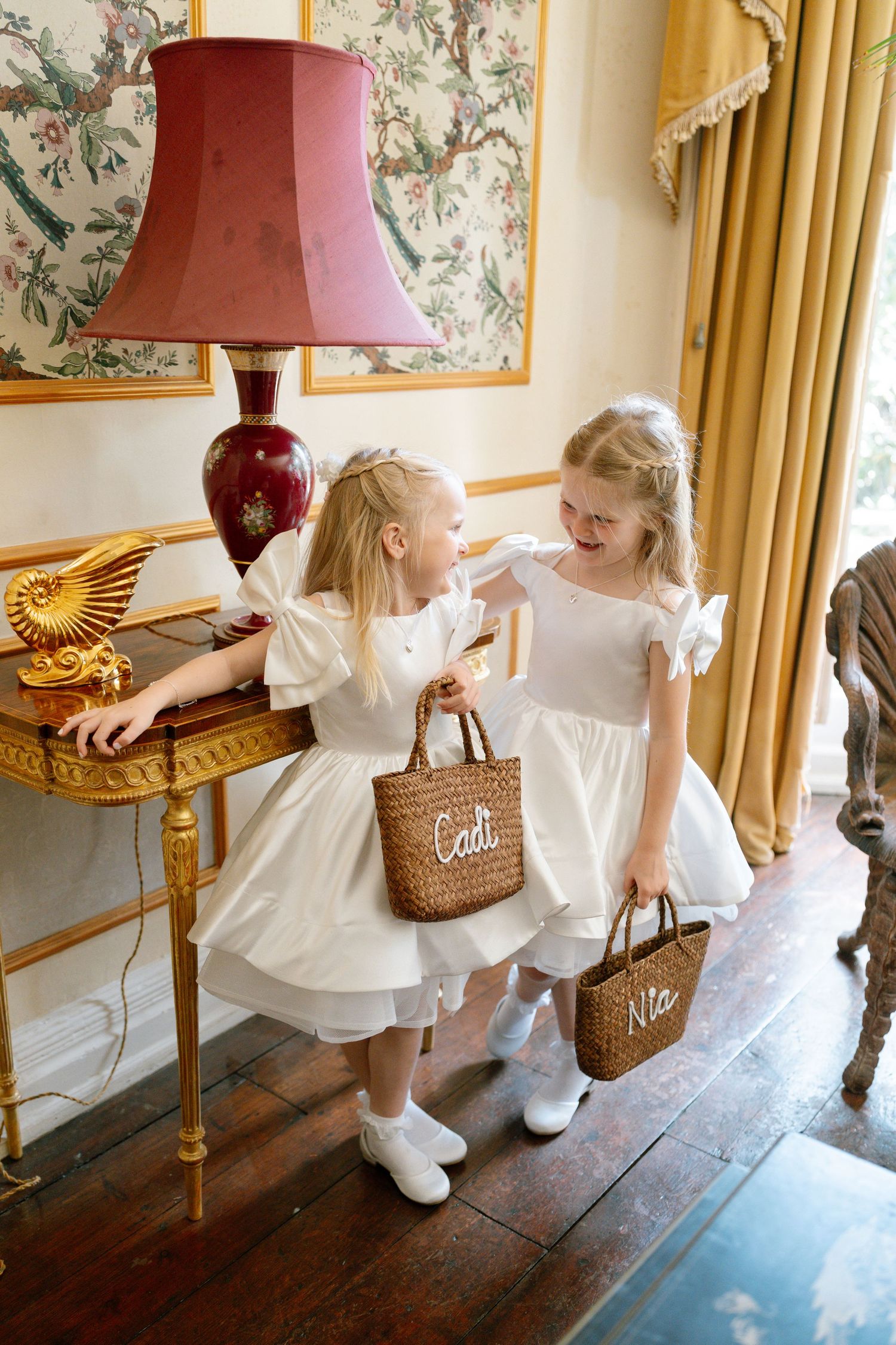 Children in white dresses play with wicker baskets in an ornate hallway.