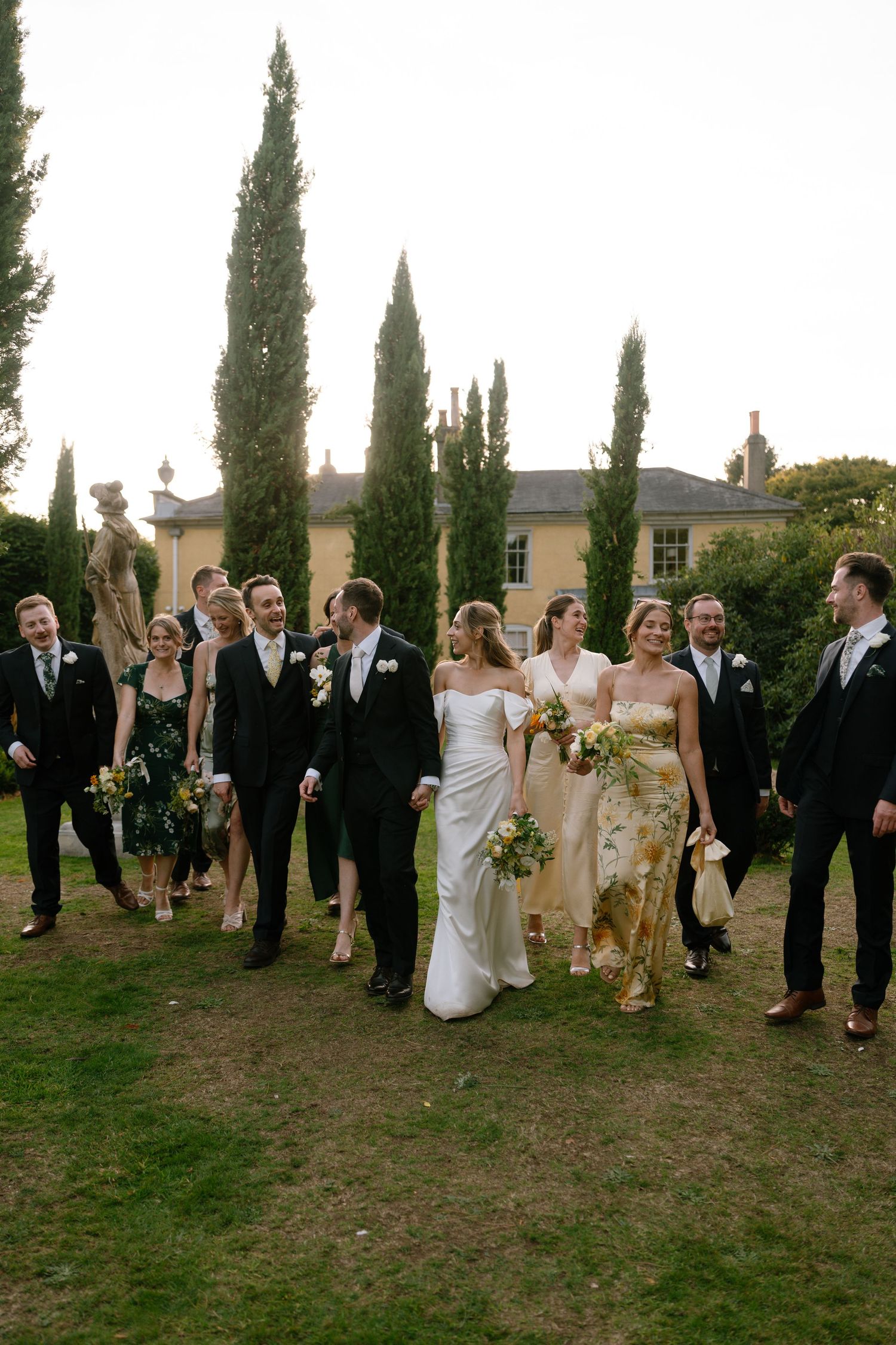 Wedding party poses for casual group shots on grass with elegant building and cypress trees in background.
