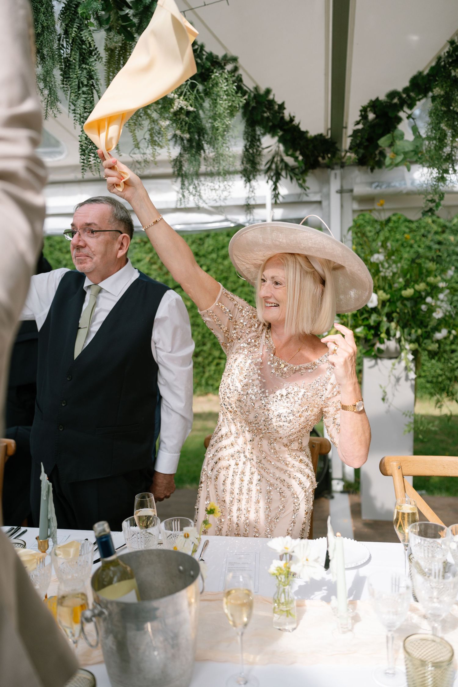 Guest in sparkly dress waving napkin during celebration under tent with hanging greenery.