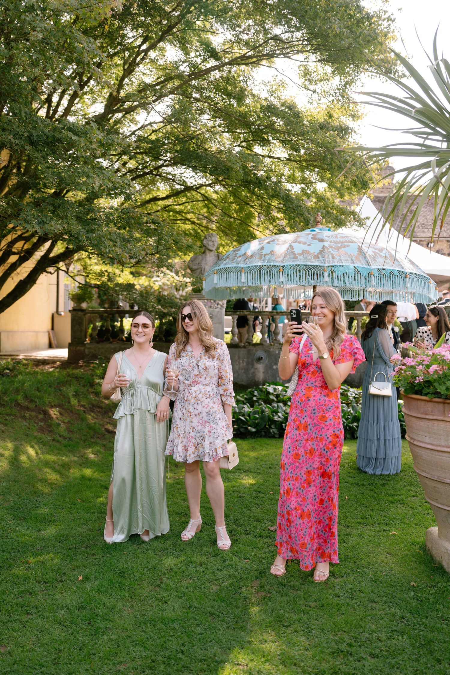 Wedding guests in colorful floral dresses gather on a grassy lawn during an outdoor celebration.