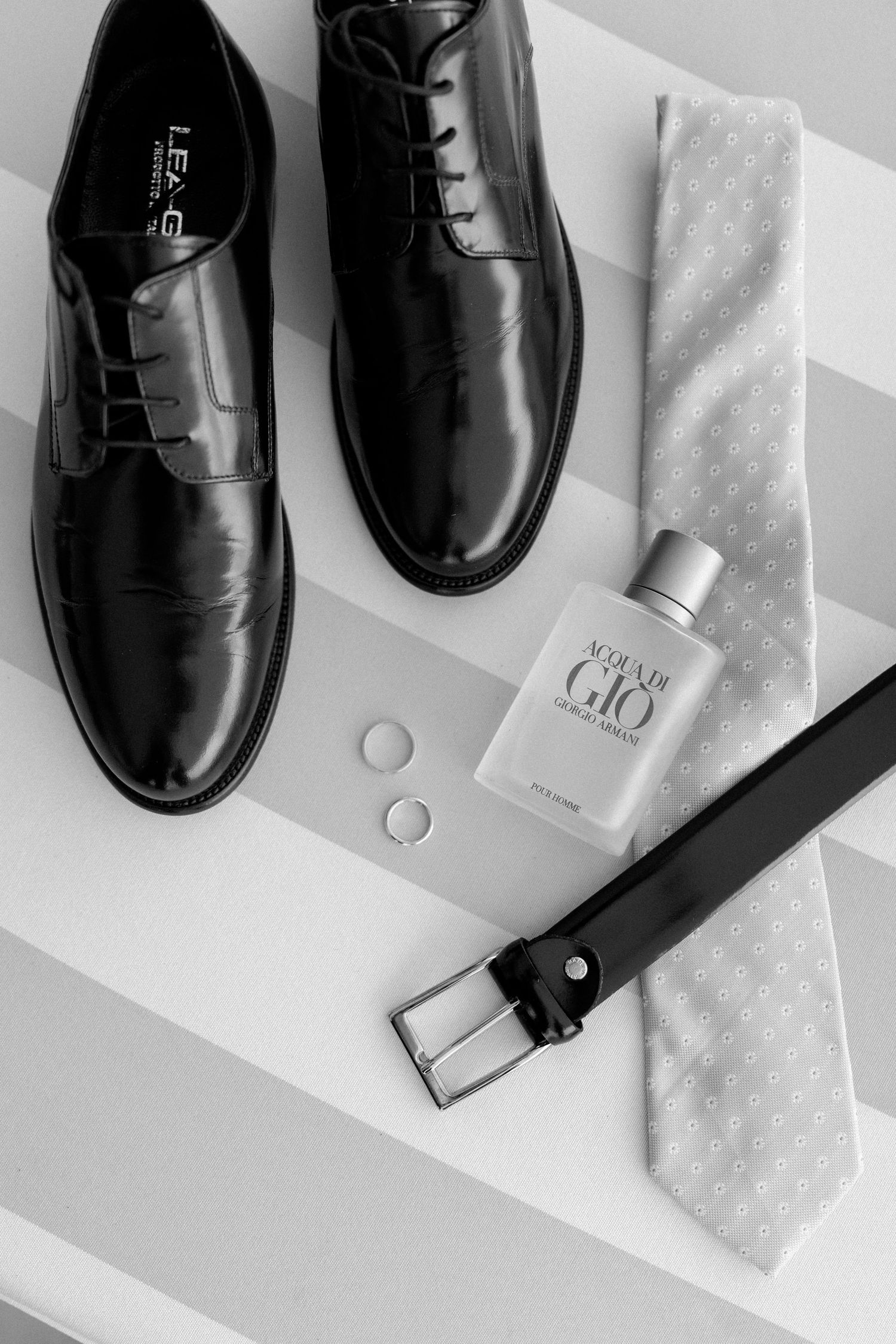 Men's formal black leather shoes with cologne, rings and tie on gray background.
