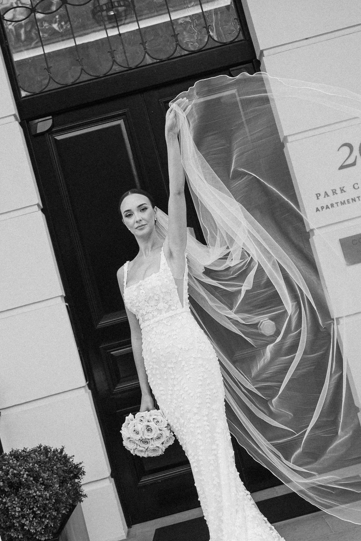 Black and white portrait of bride with flowing veil and bouquet by elegant doorway.