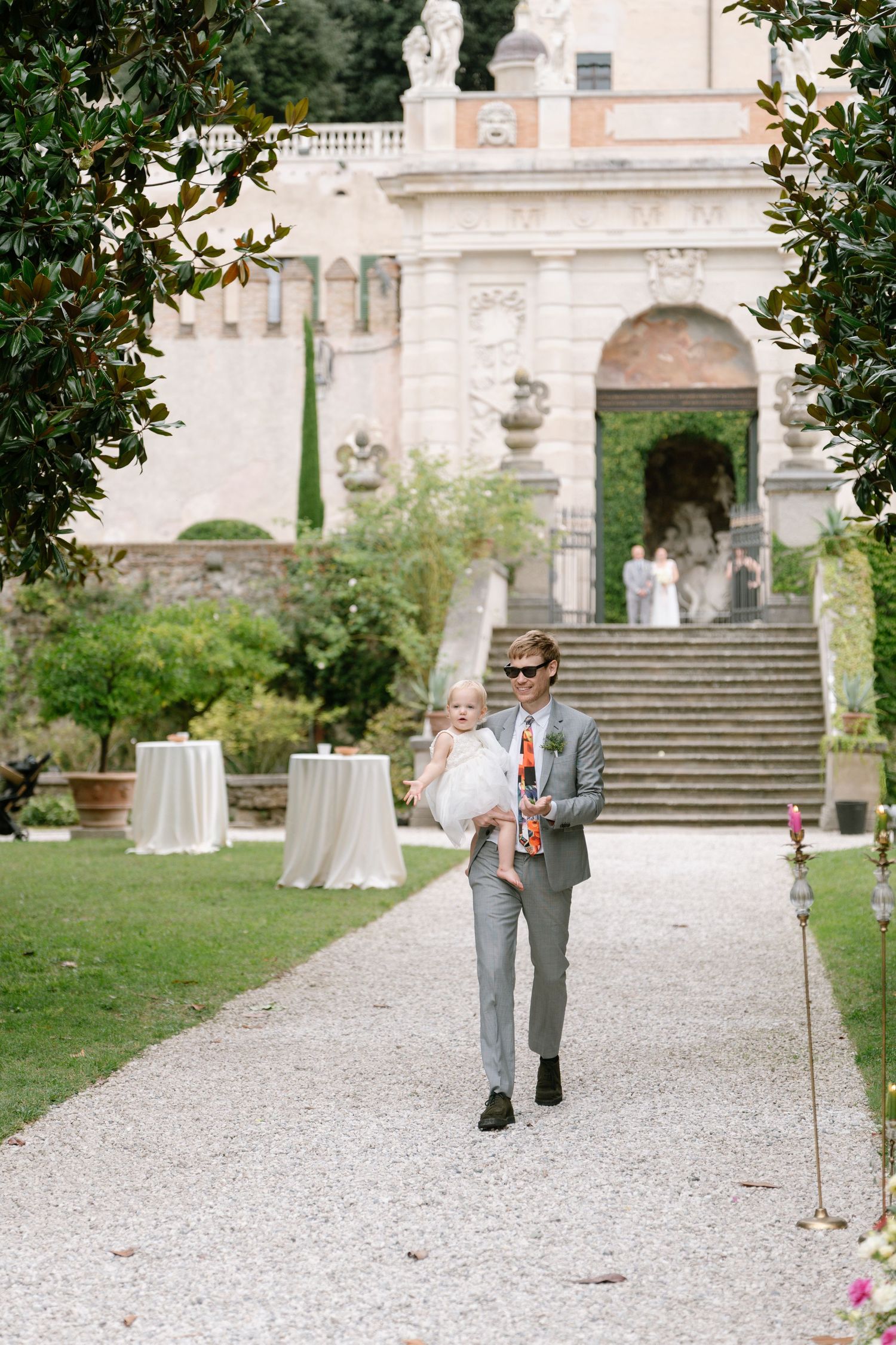 People walking along a gravel path leading to grand stone steps of an Italian villa.