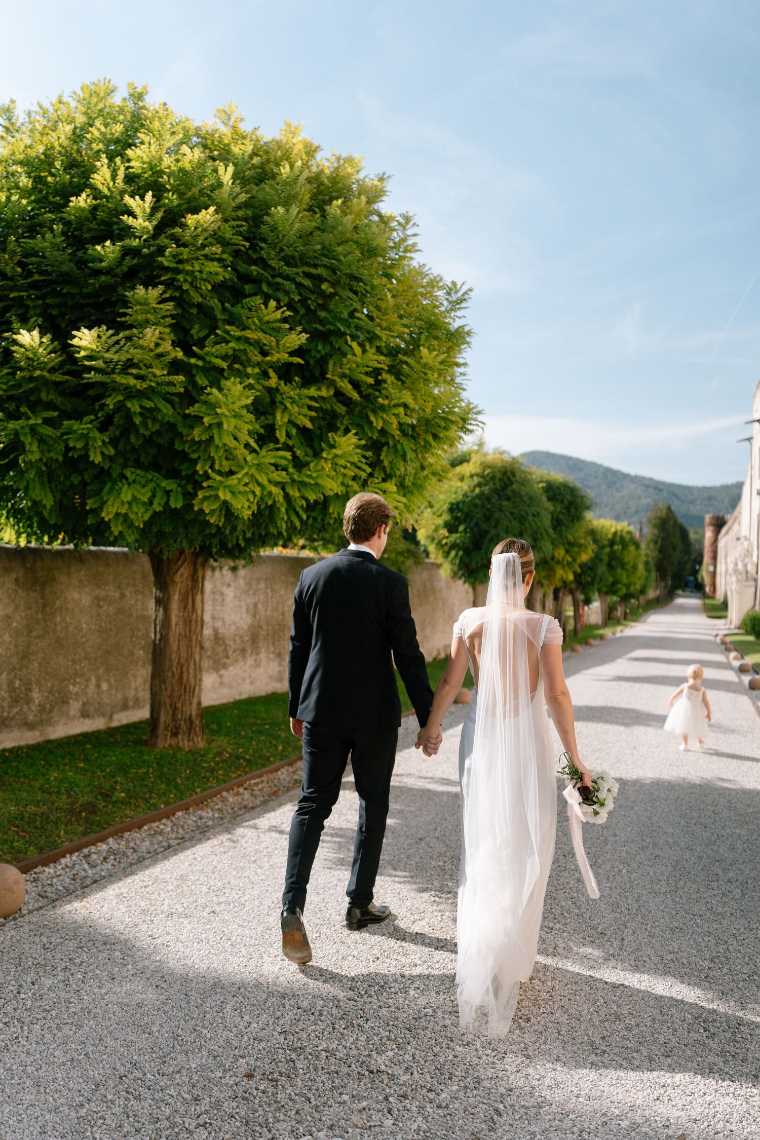 Newlyweds walking down tree-lined path holding hands on sunny wedding day.
