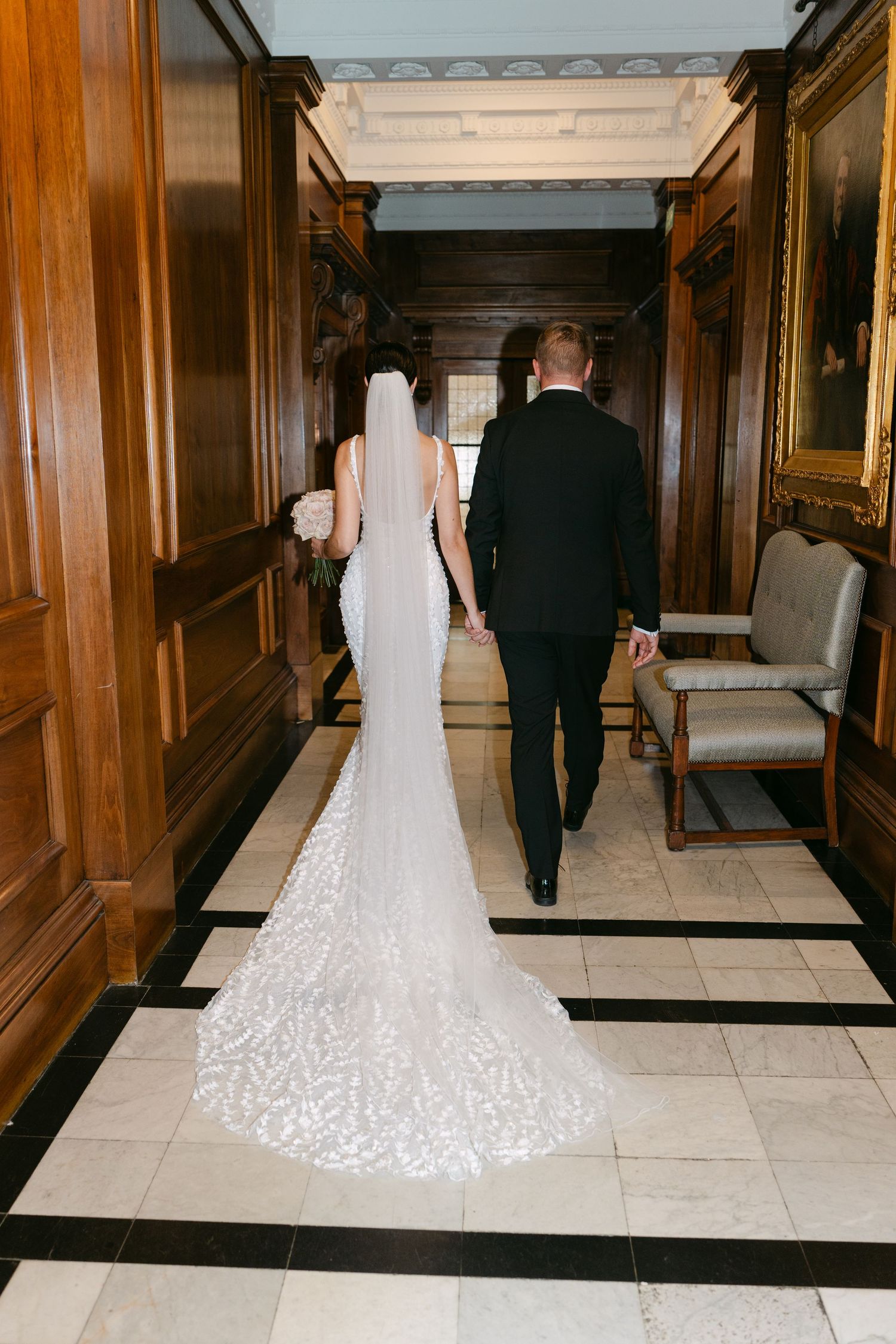 Bride and groom walk down elegant hallway showing detailed train of lace wedding dress.