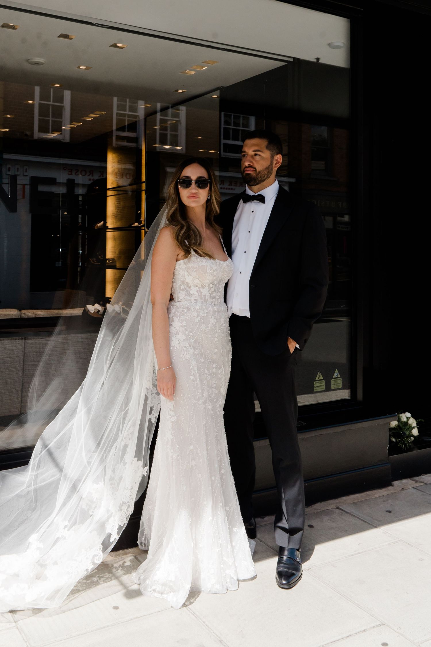 A bride in sunglasses and groom in tuxedo walk together in an elegant indoor setting.