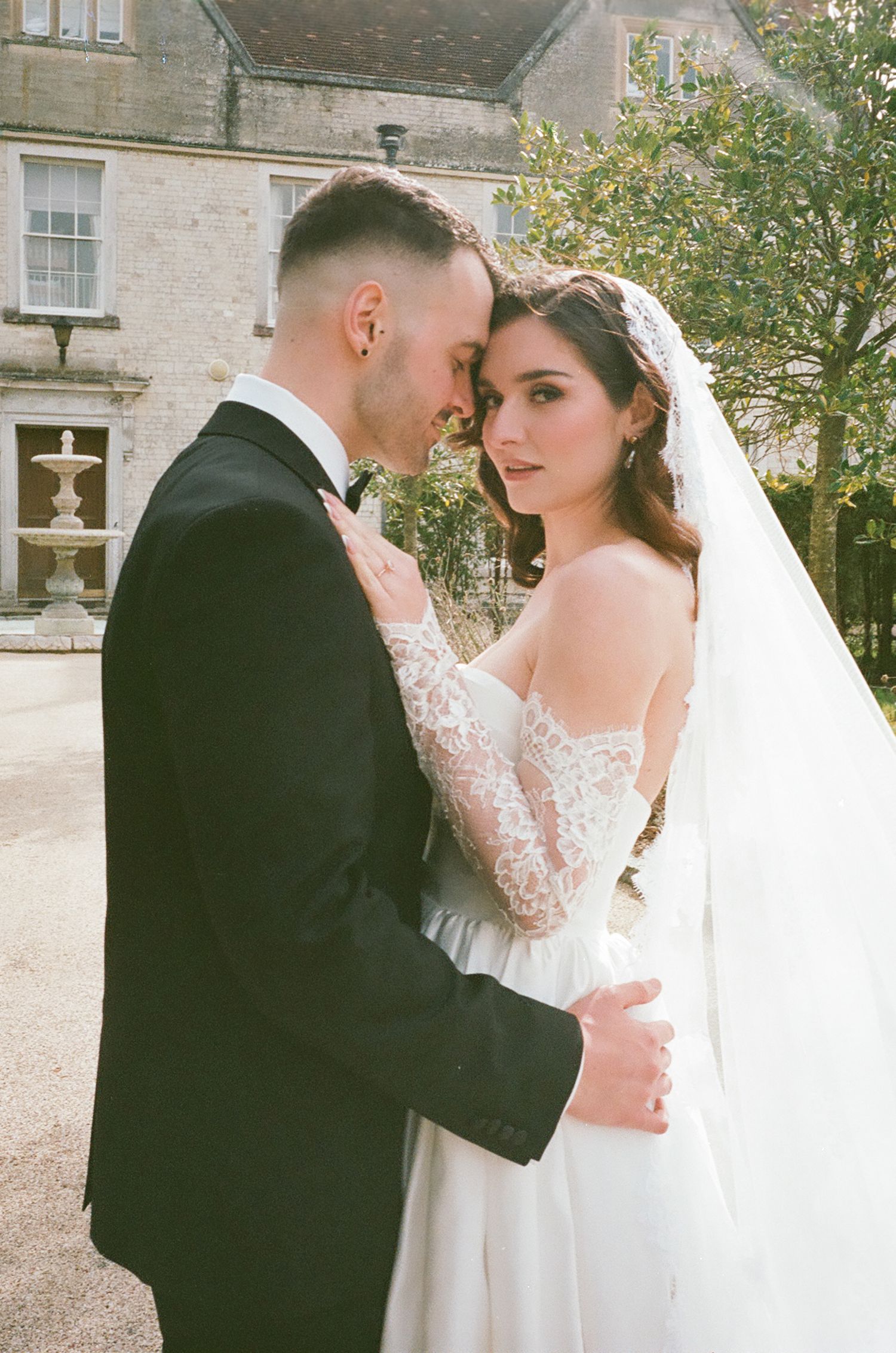 A romantic wedding photo of a couple embracing outdoors in front of a stone building with a lace gown and black tuxedo.