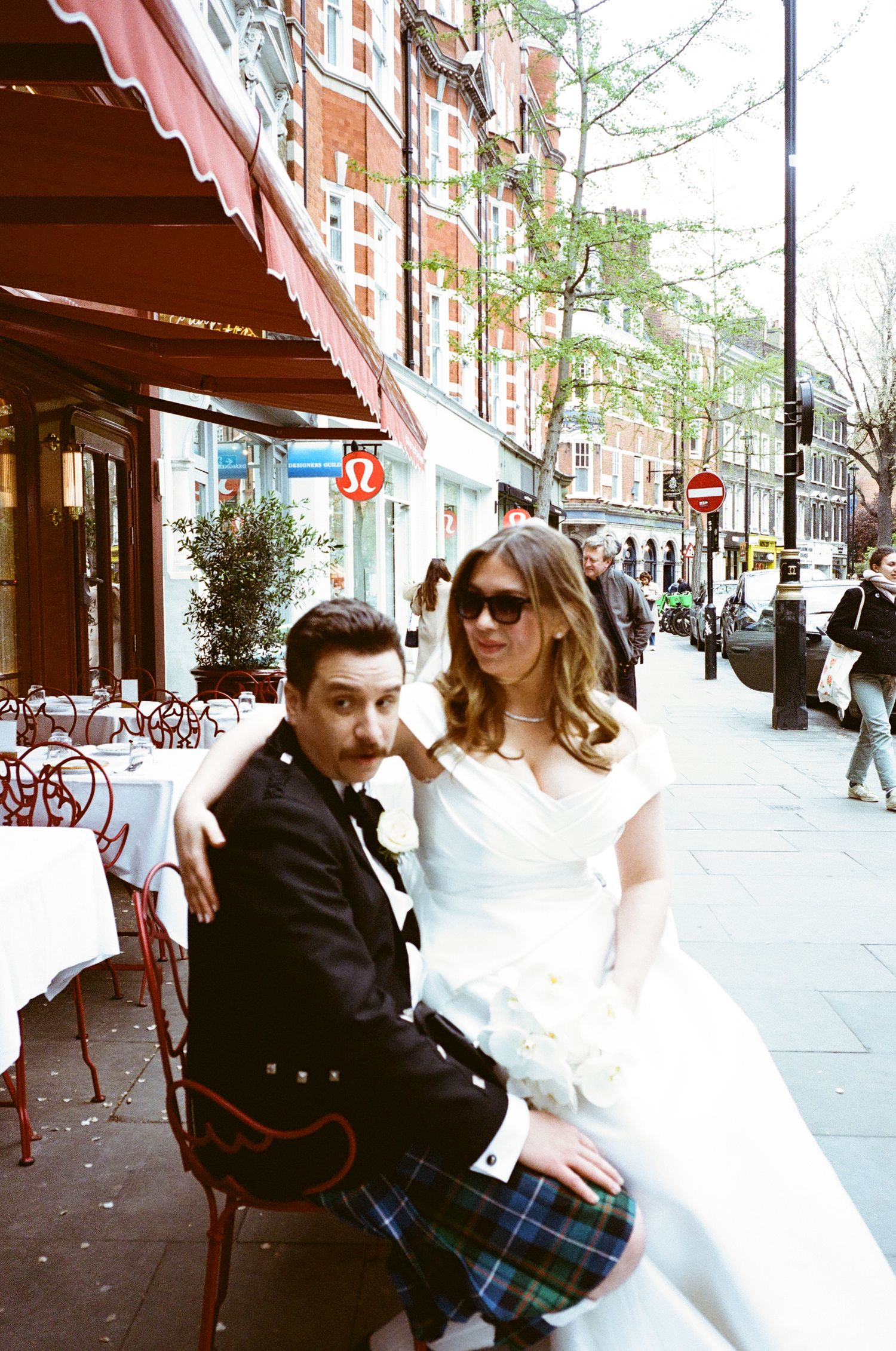 A couple sits together on a bench outside a storefront on a city sidewalk with red awnings and trees.