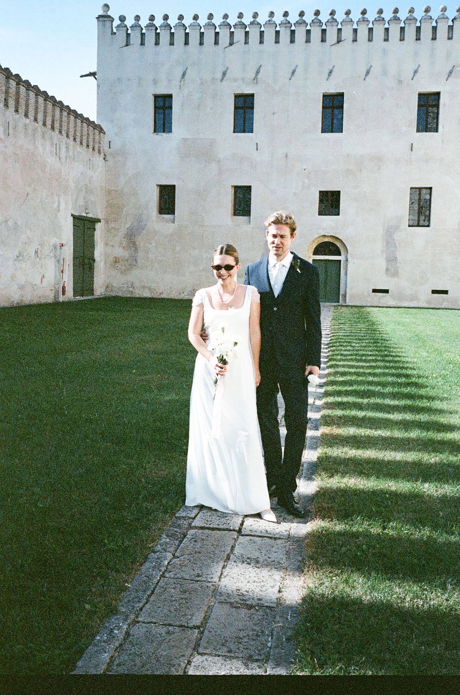 A couple in wedding attire stands on a stone path beside a white historic building with green trim and ornate details.