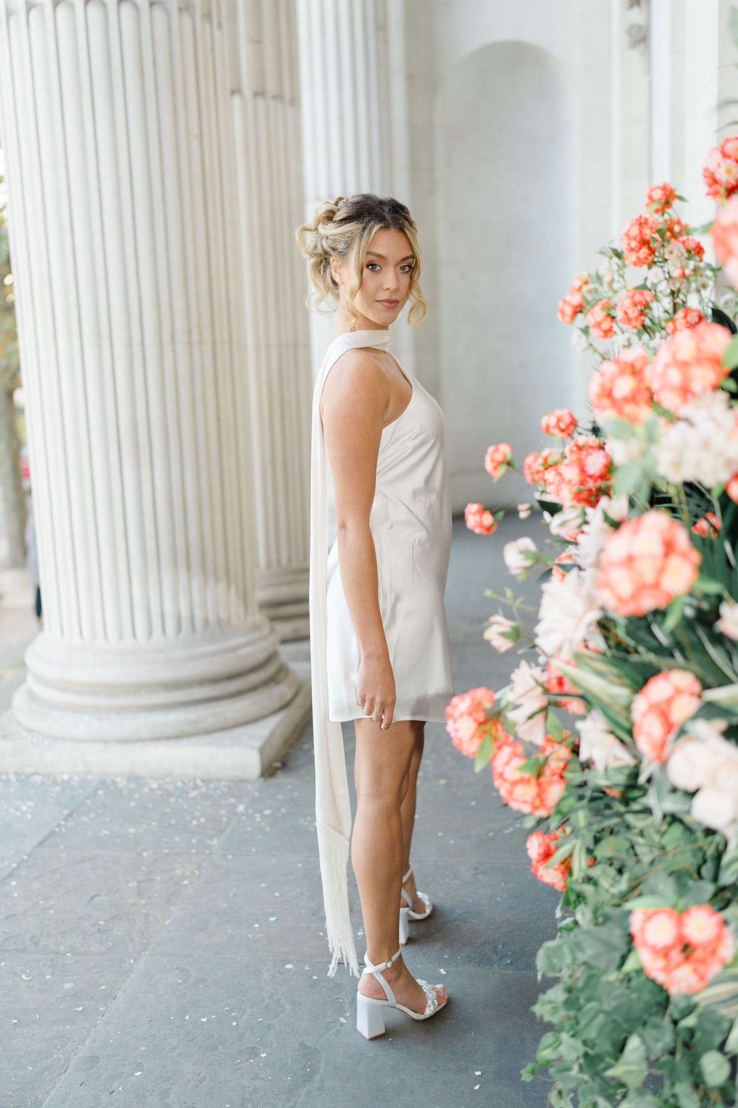 Woman in white dress poses elegantly among columns with coral flowers.