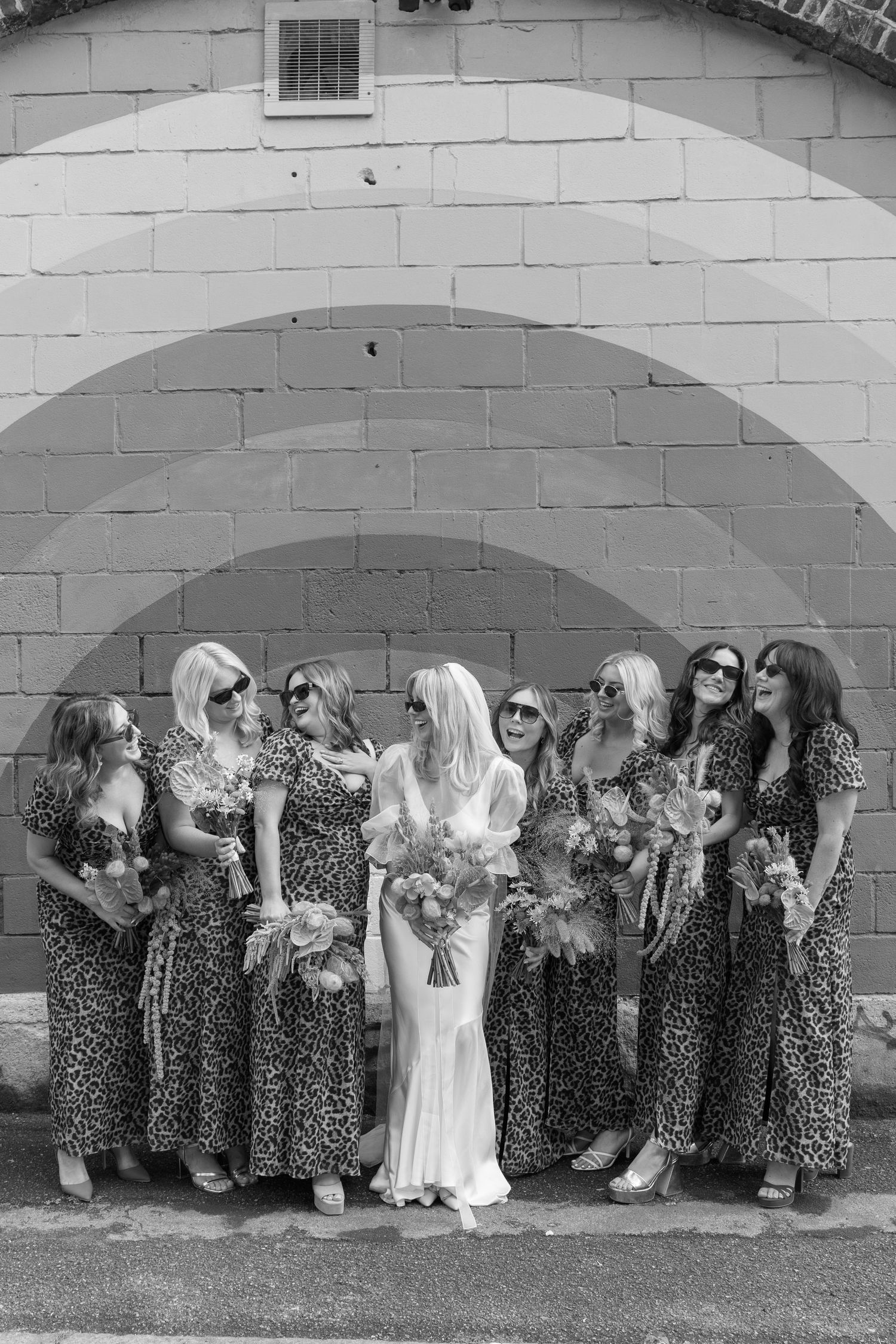 Wedding party poses against white wall wearing matching leopard print dresses in black and white.
