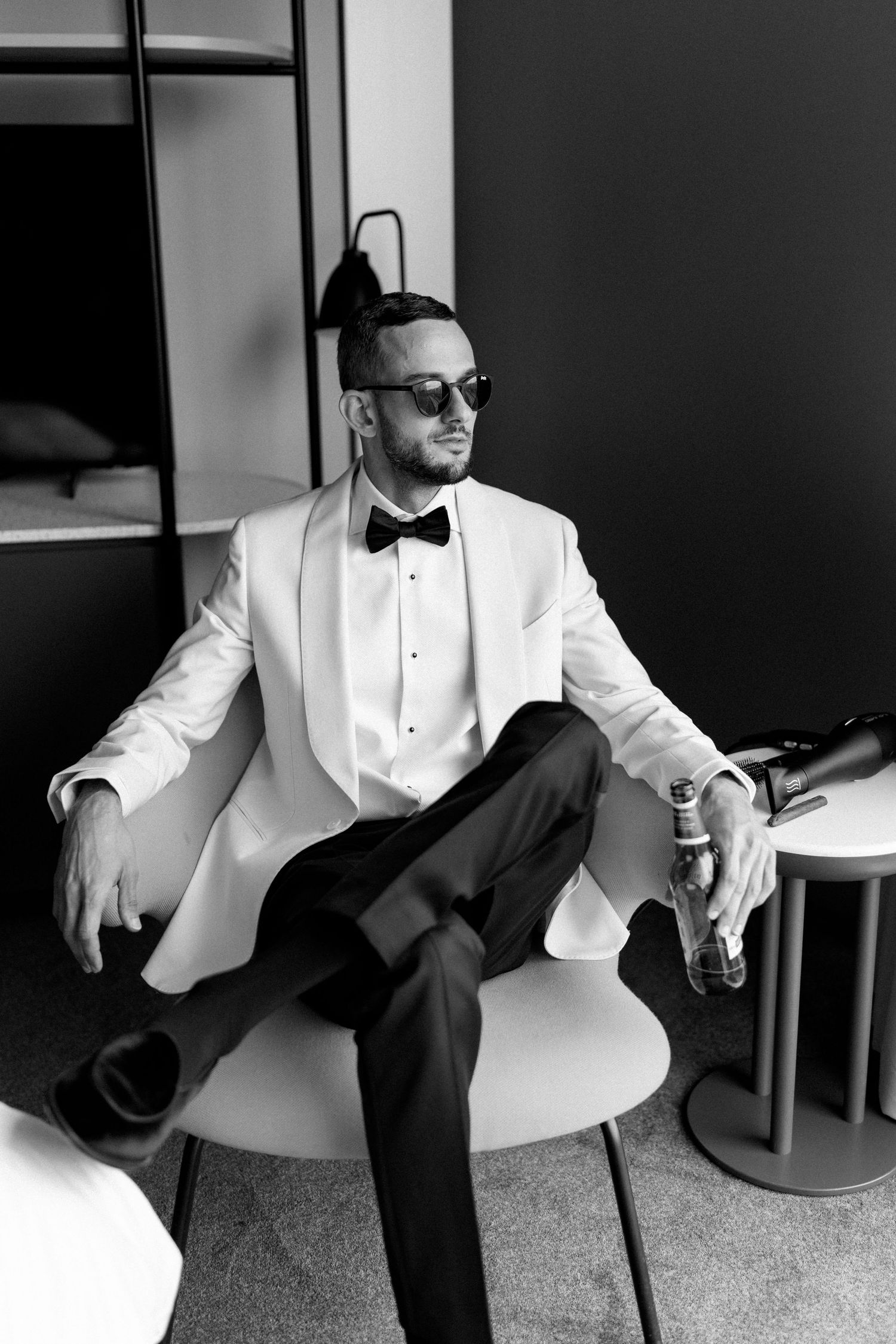 Formal portrait of person in white tuxedo jacket and sunglasses seated in modern chair.