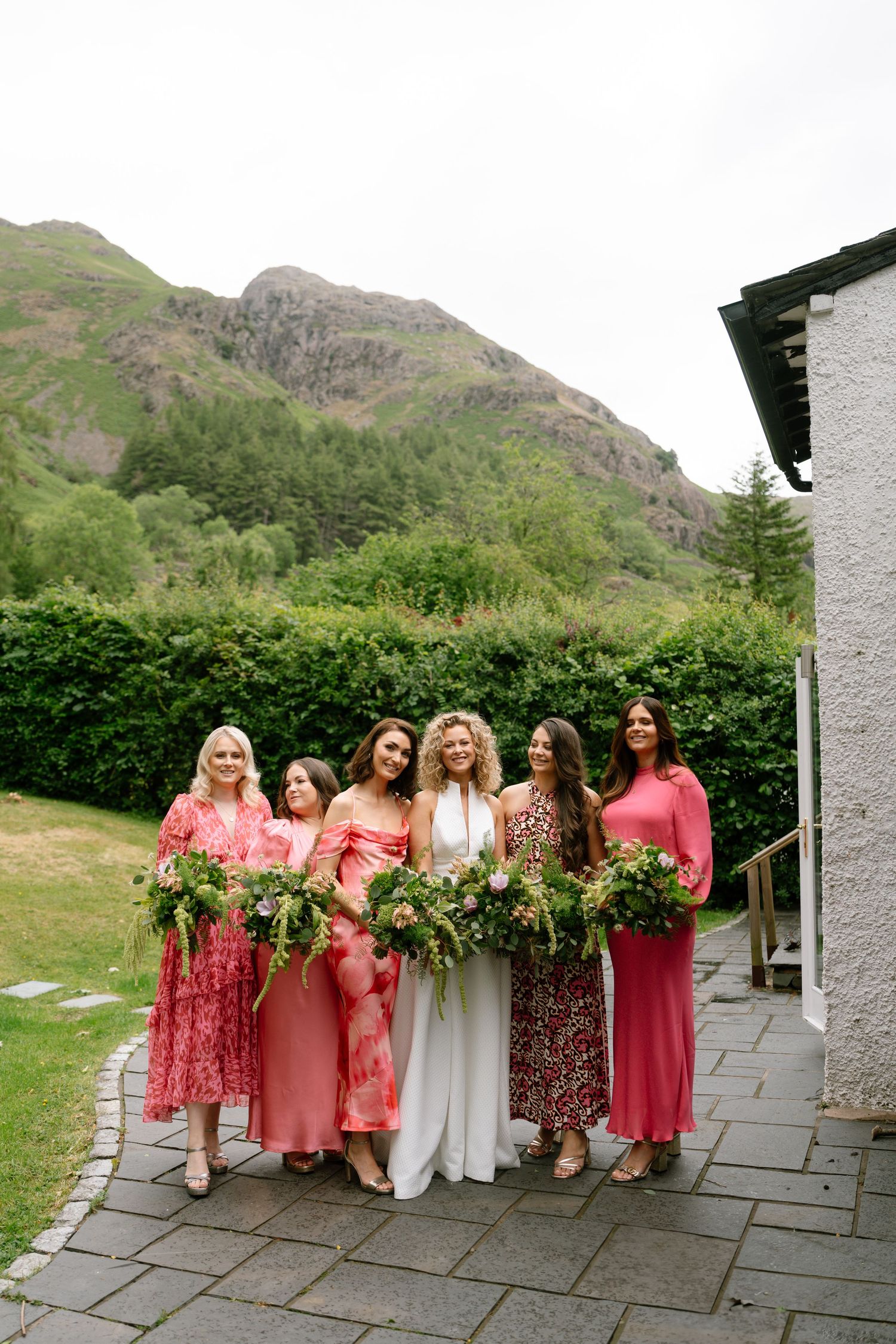 Bridesmaids in coral and floral dresses pose together against a mountain backdrop.