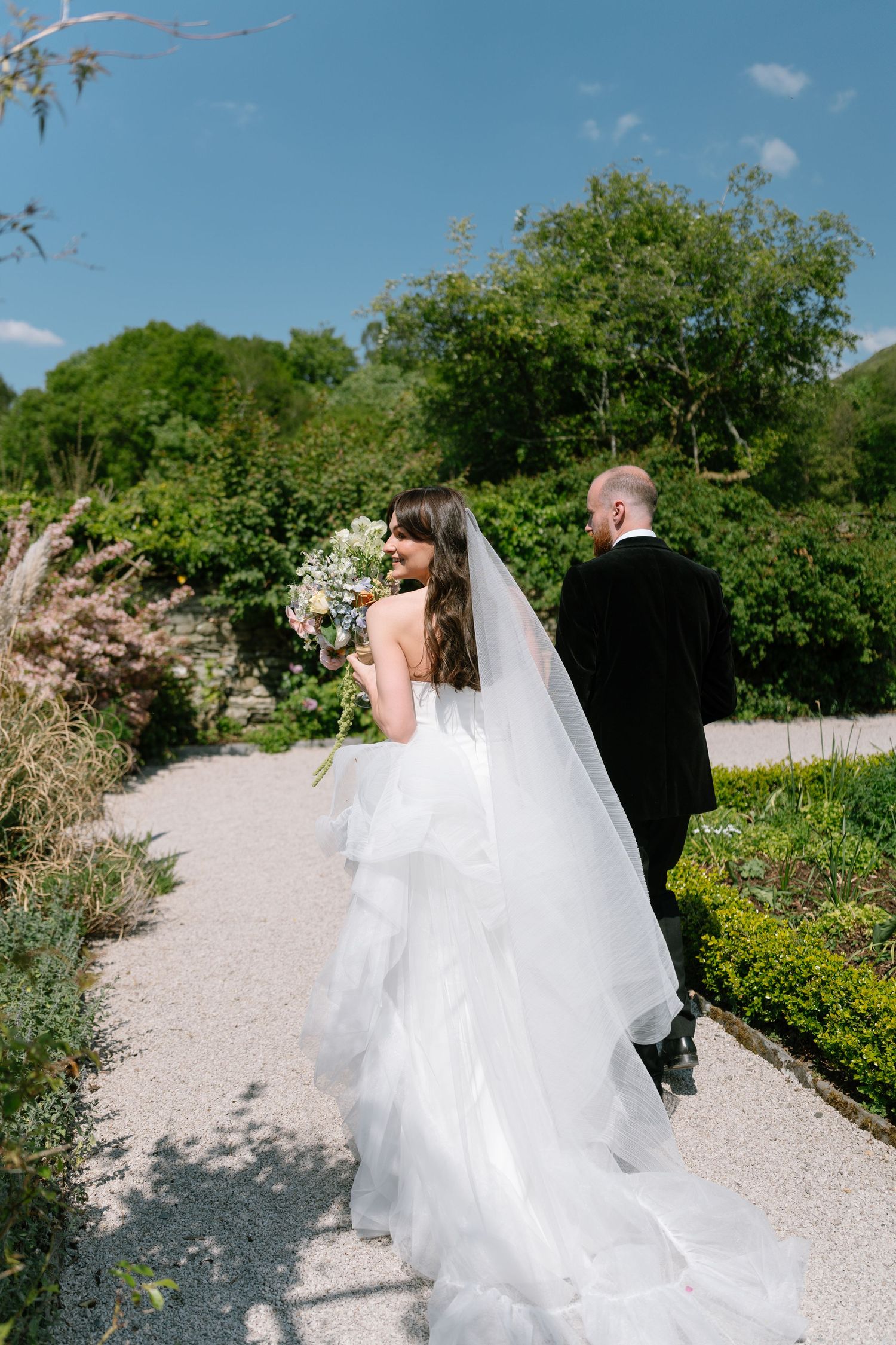 A wedding couple walks along a gravel garden path surrounded by lush greenery on a sunny day.