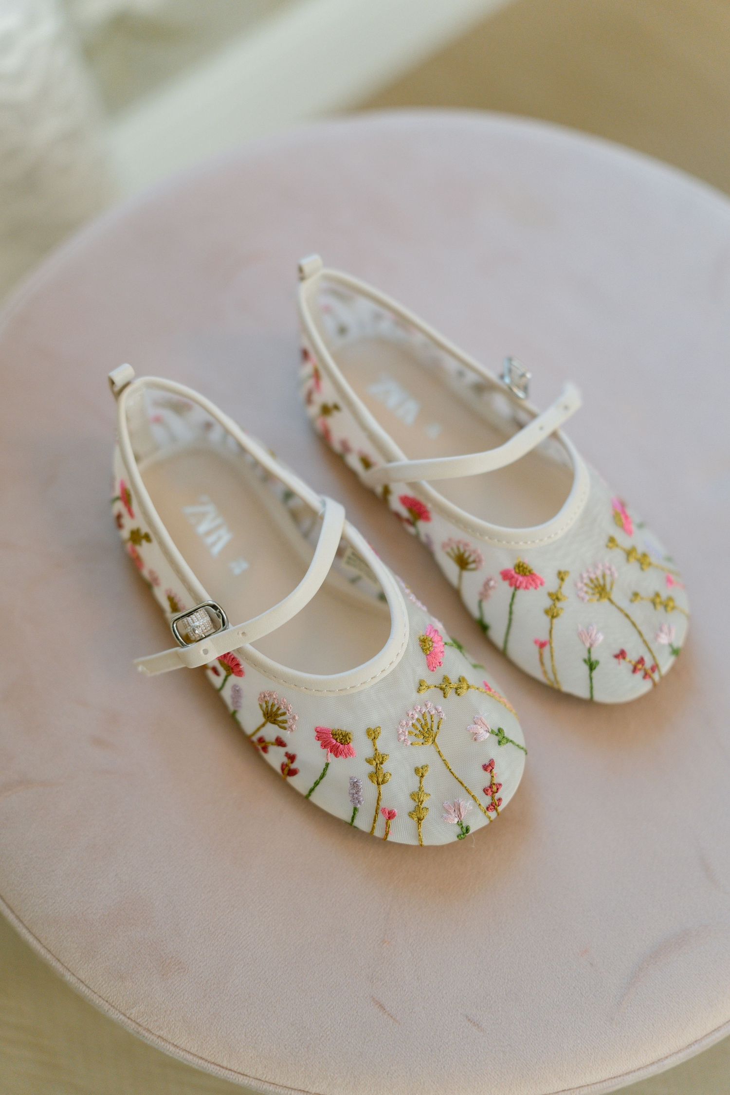 White baby shoes with delicate floral embroidery displayed on a pink surface.