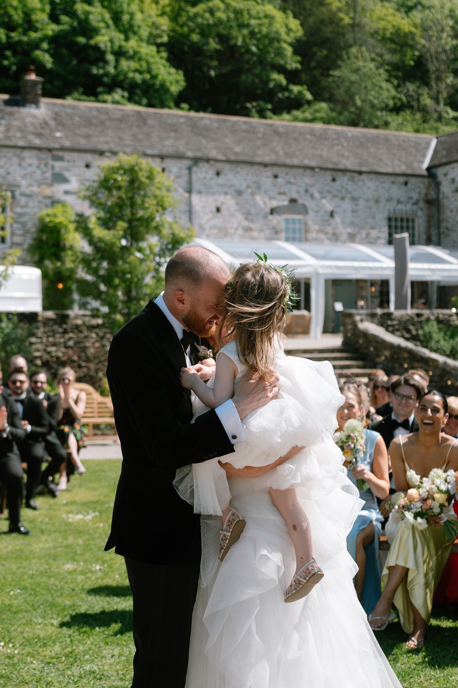 A romantic outdoor wedding ceremony shows the couple sharing their first kiss in front of stone buildings and guests.