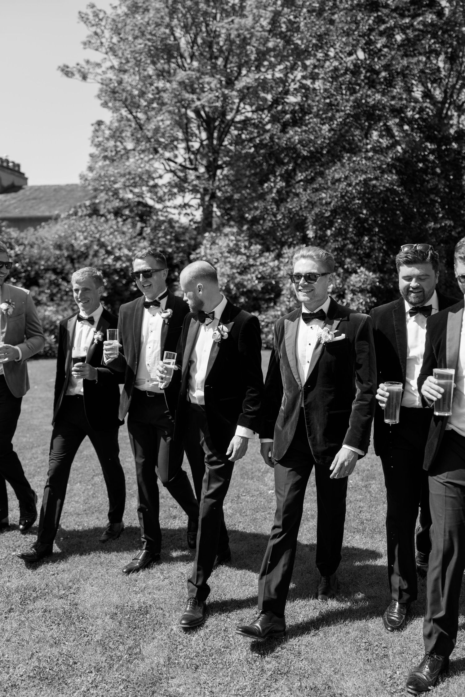 Wedding party members in tuxedos pose casually on grass while holding drinks.