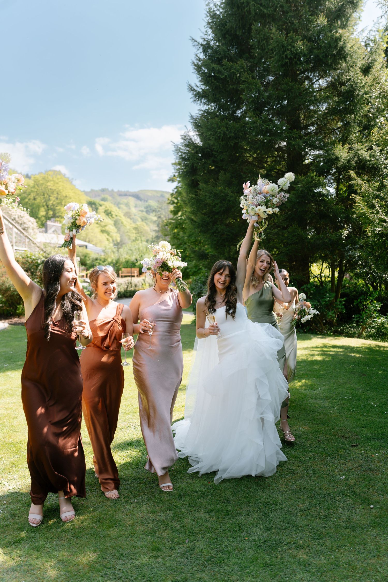 Joyful wedding party in brown and neutral dresses dances on grass during outdoor photo session.