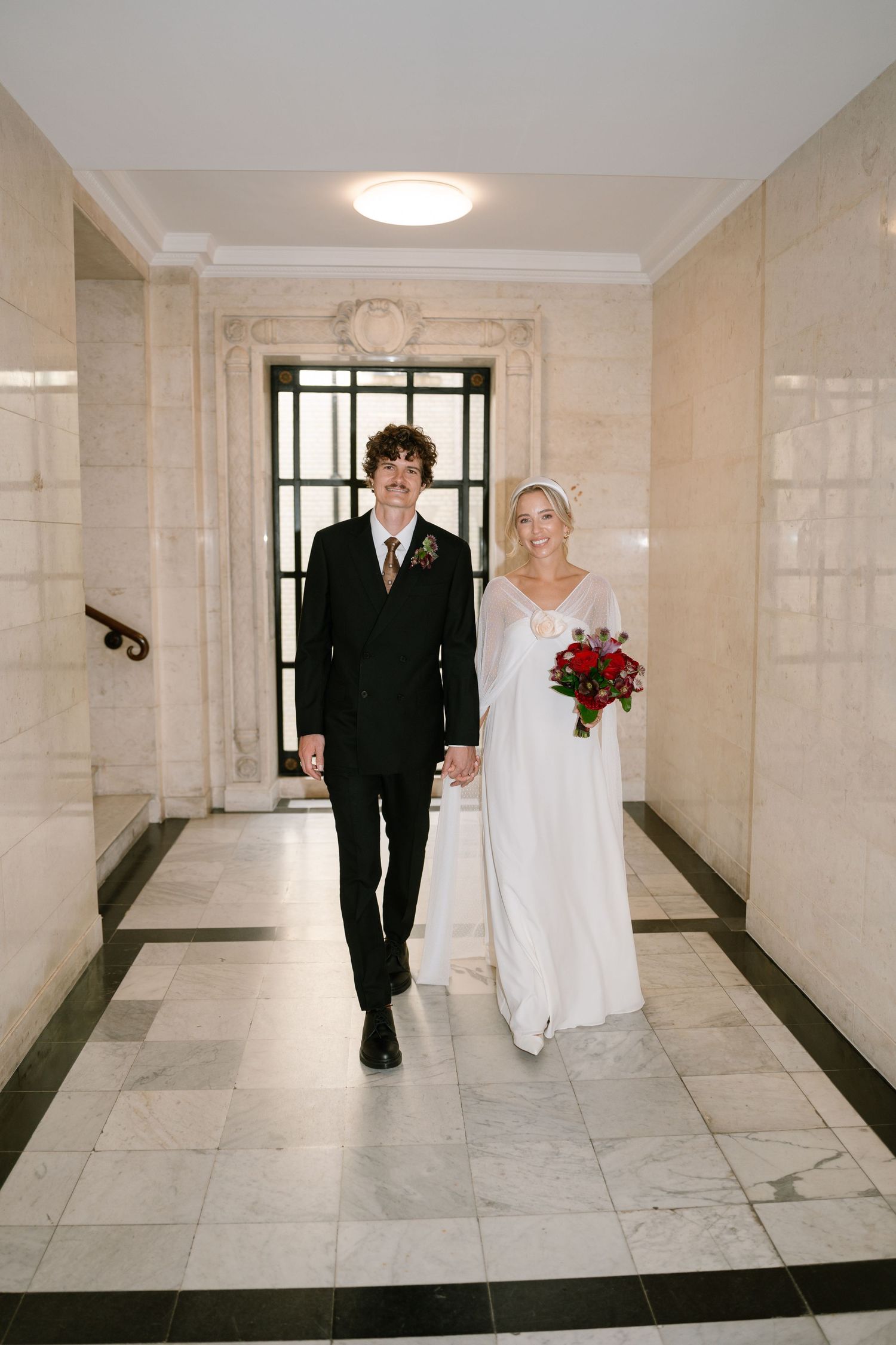 Bride and groom walking through an elegant marble corridor with natural lighting.