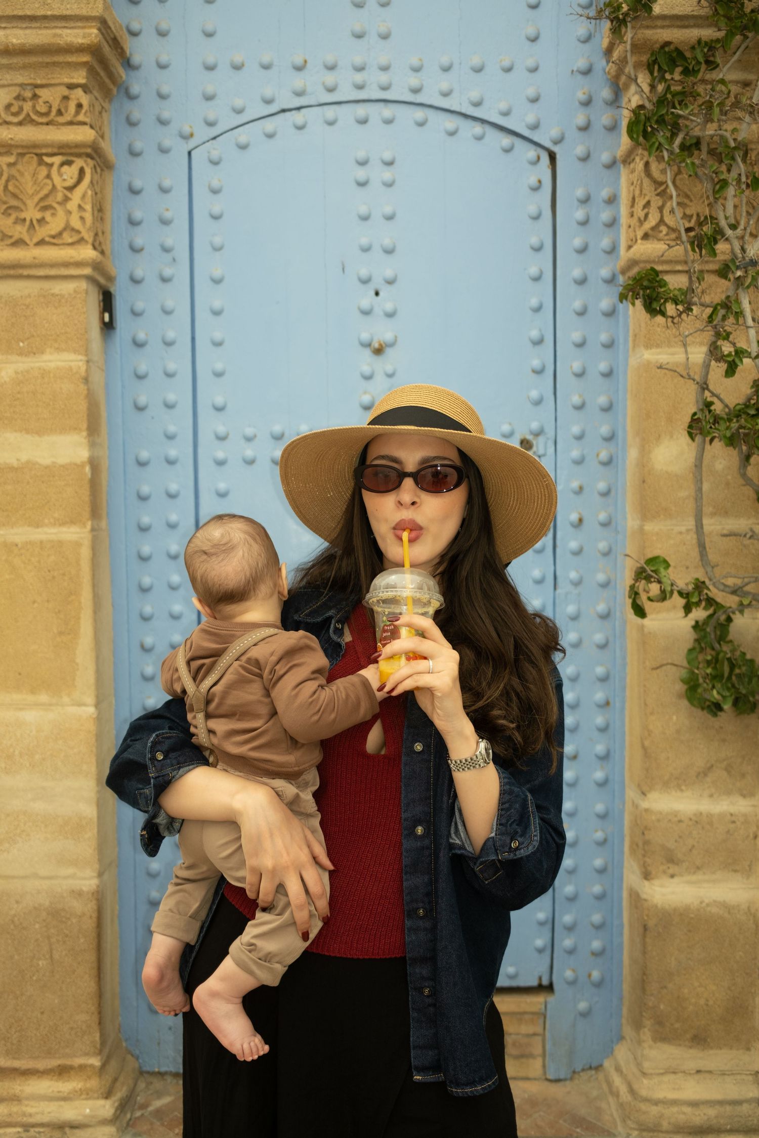 Woman in straw hat and sunglasses sipping iced drink while holding a baby in front of blue door.