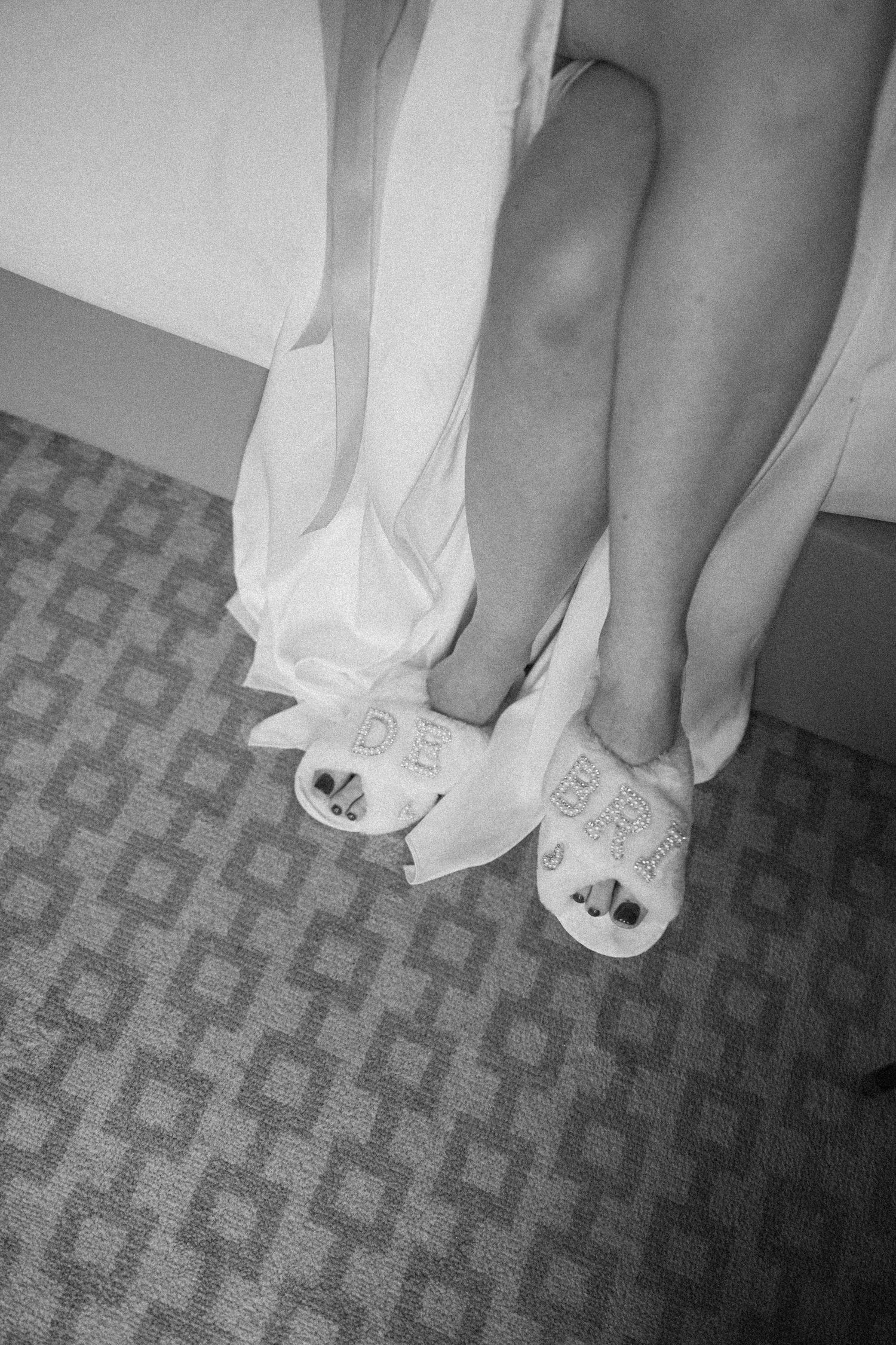 Black and white photo of a bride wearing feathered wedding dress and bride slippers on patterned carpet.
