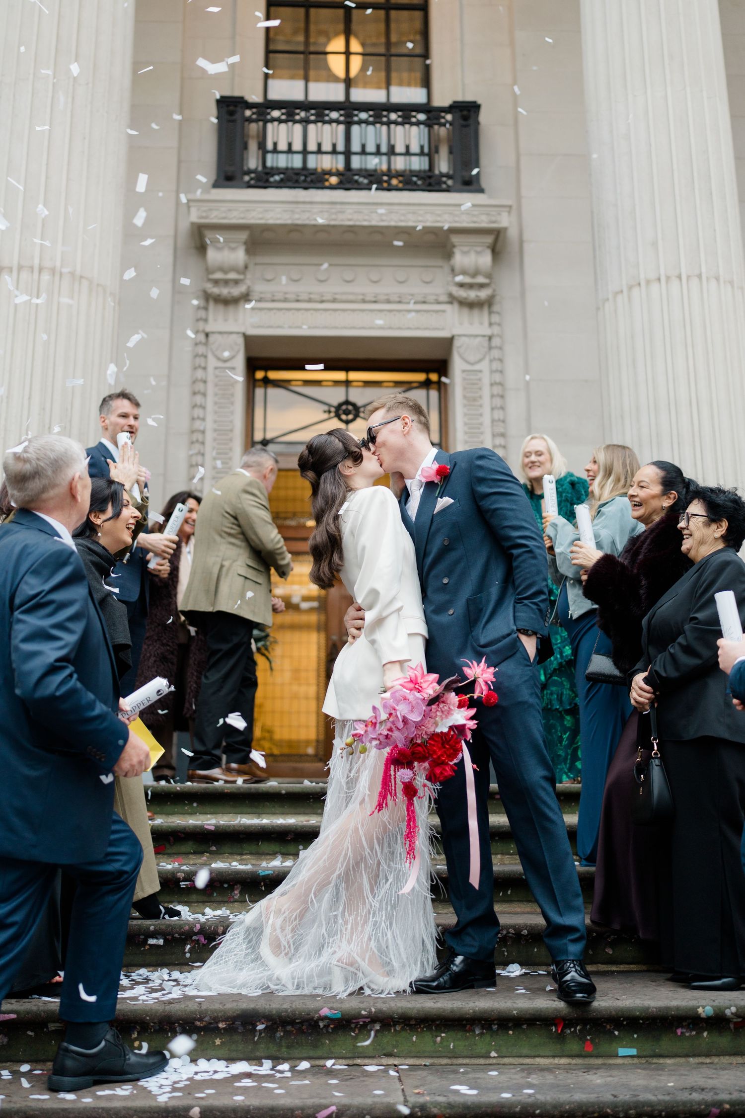 Newlyweds share a kiss on steps as guests throw white confetti outside a grand neoclassical building.