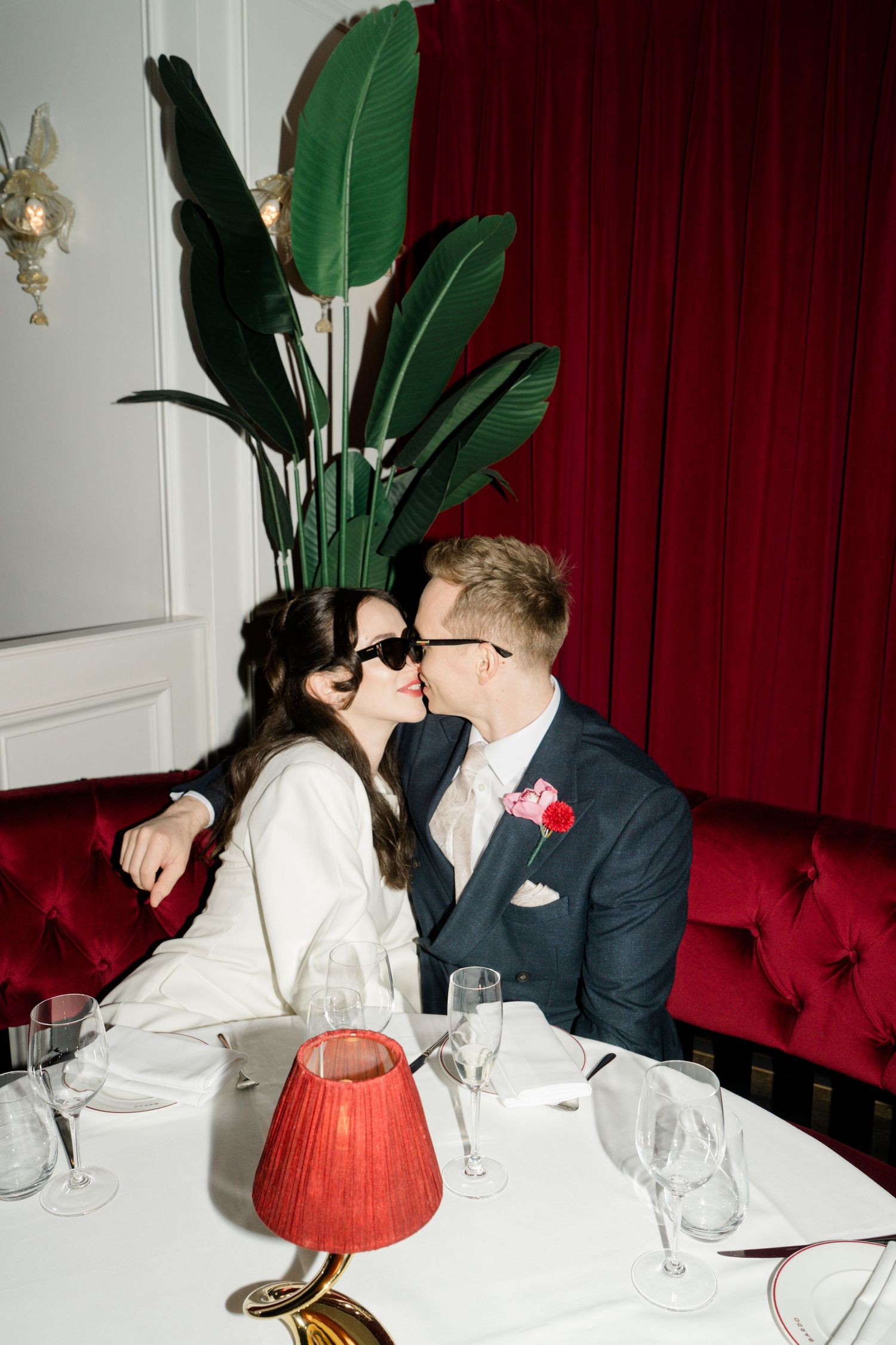 Bride and groom kissing at elegant restaurant table with red velvet booth and tropical plant.