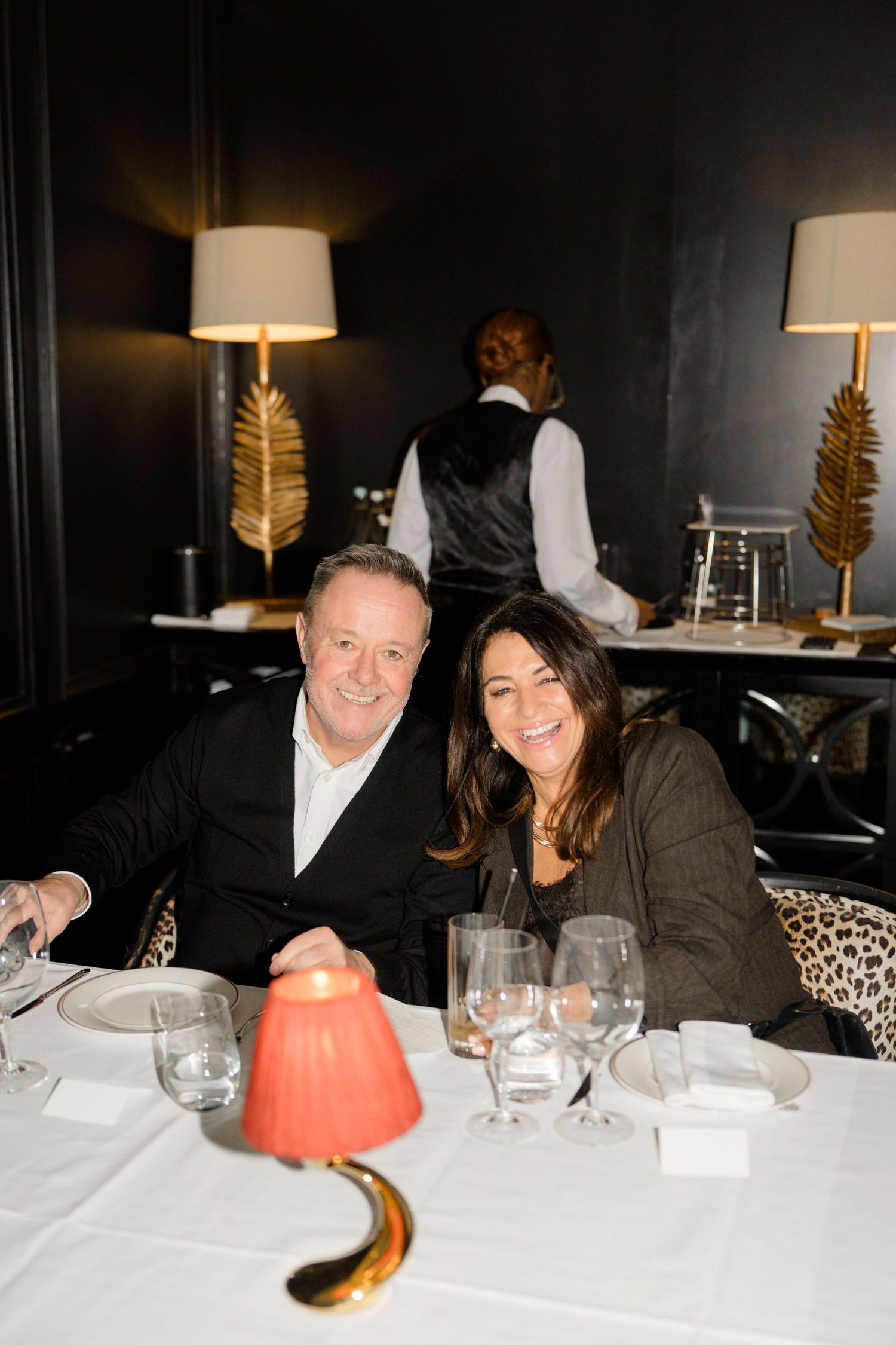Smiling couple seated at elegant restaurant table with white tablecloth and gold leaf lamp decor.