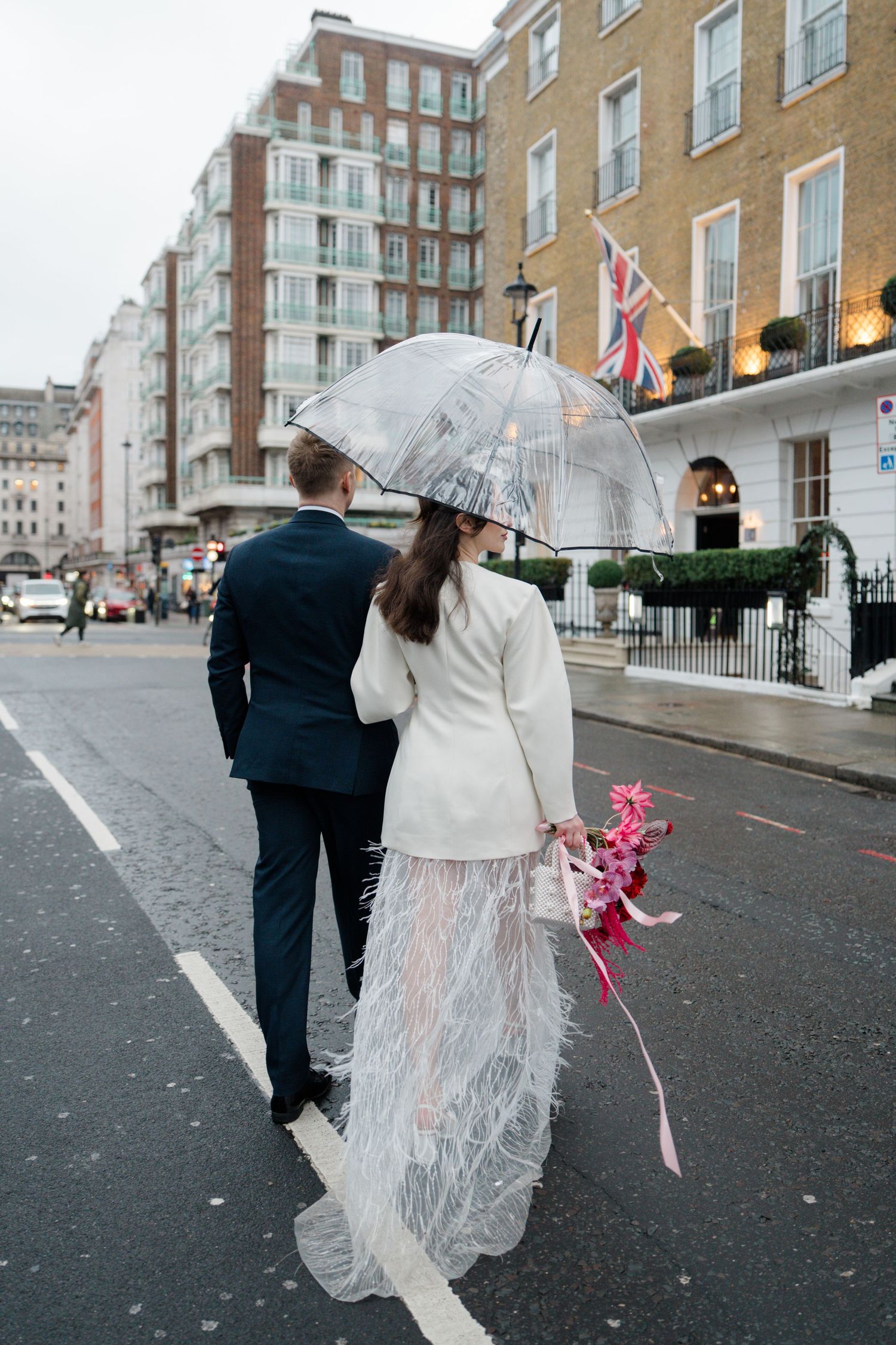 Bride and groom walking street in London under clear umbrella on rainy wedding day.