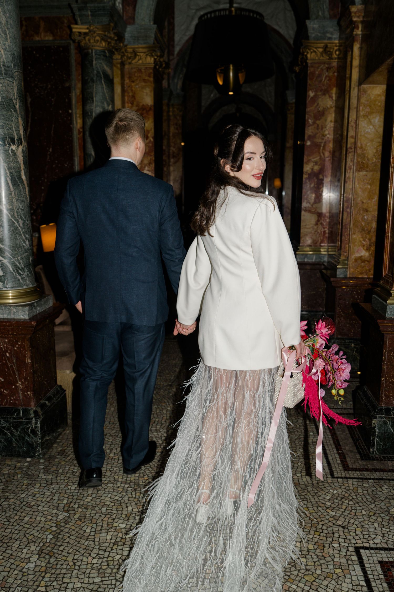 Bride in white blazer and feather skirt holds pink bouquet walking with groom in navy suit.