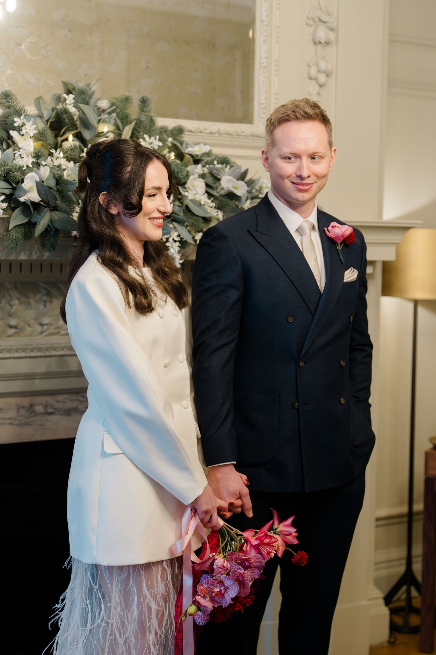 Bride in white feather coat dress and groom in navy suit hold hands at elegant ceremony.