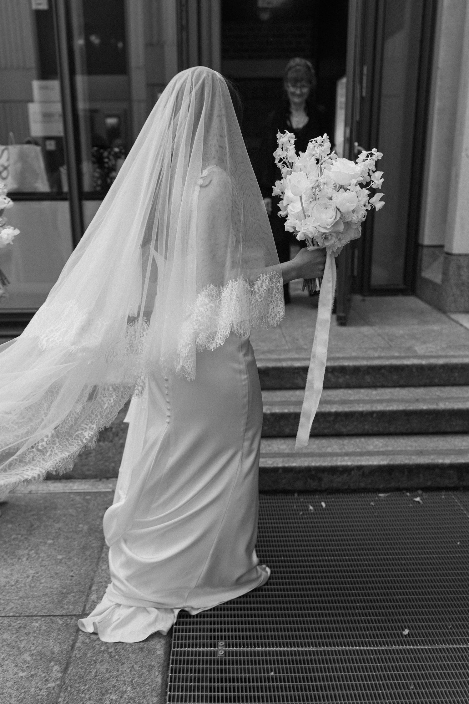 Bride in elegant satin gown and lace veil holds floral bouquet on church steps in black and white.
