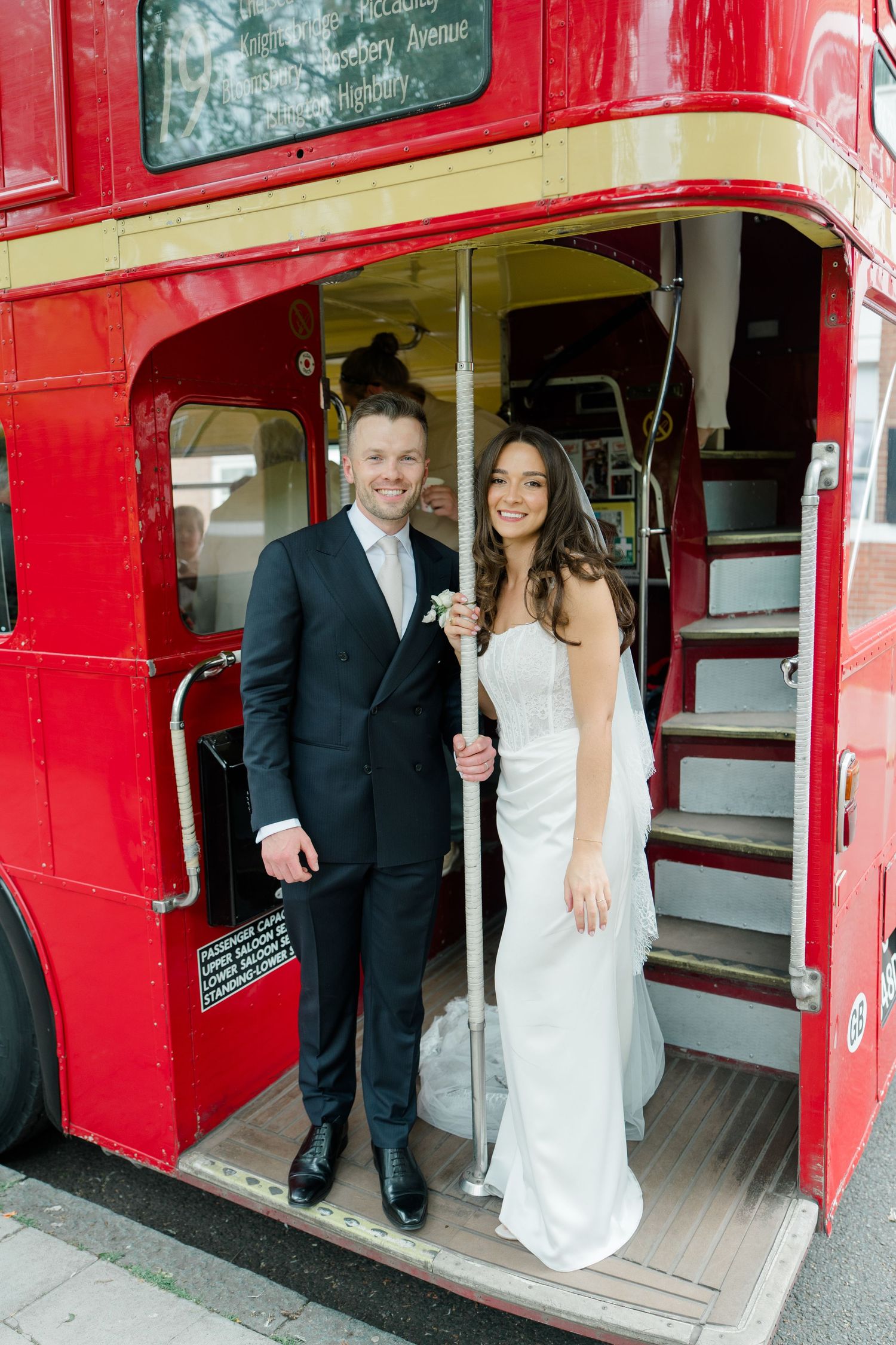 Bride and groom smiling on the steps of a classic red double-decker bus on their wedding day.