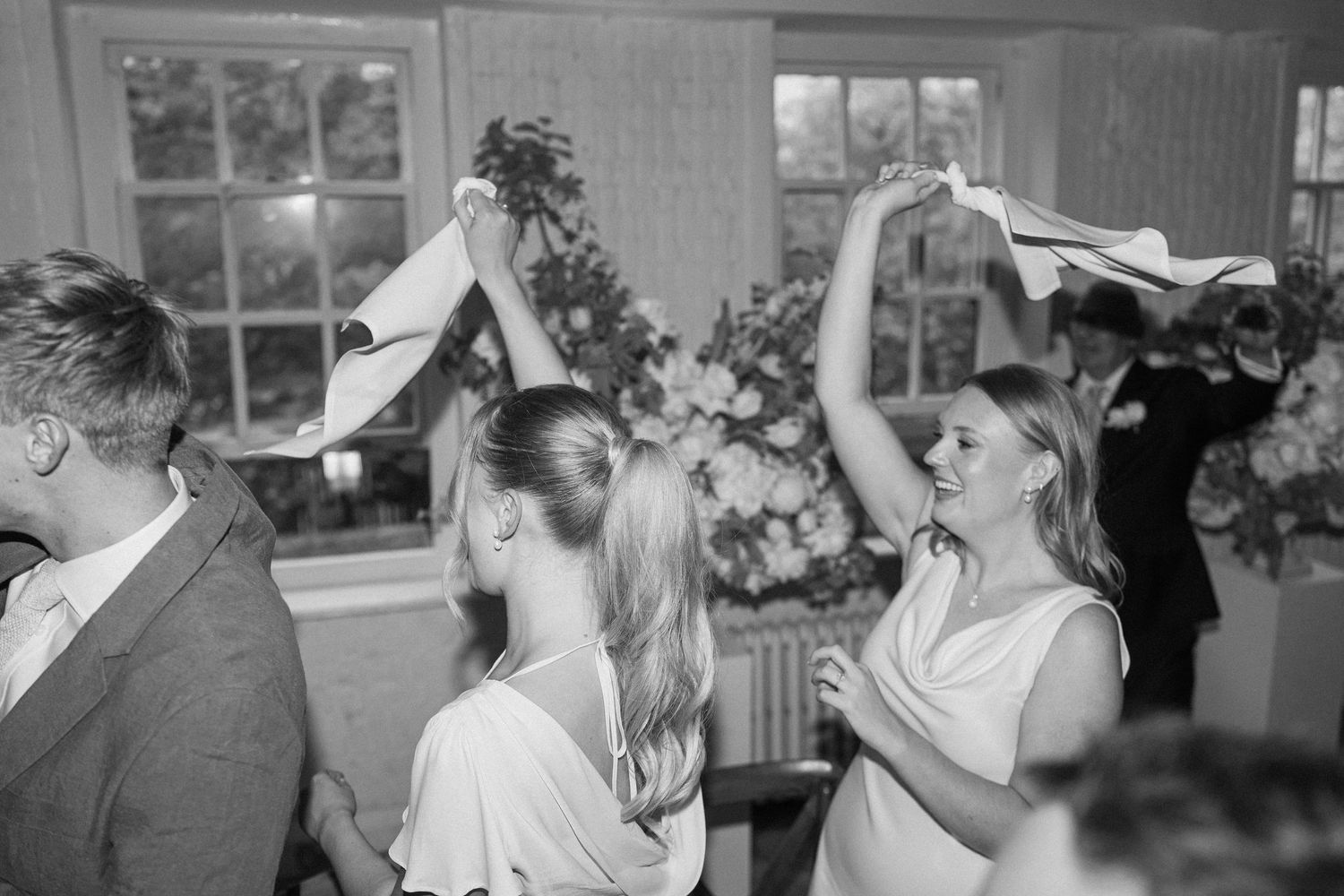 Black and white photo of wedding guests dancing joyfully at lively reception with floral backdrop.