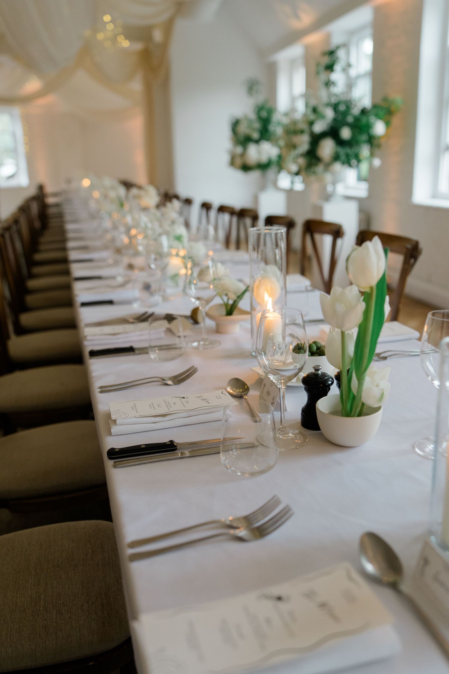 Long wedding reception table set with white tulips, candles, glassware, and neatly arranged cutlery.